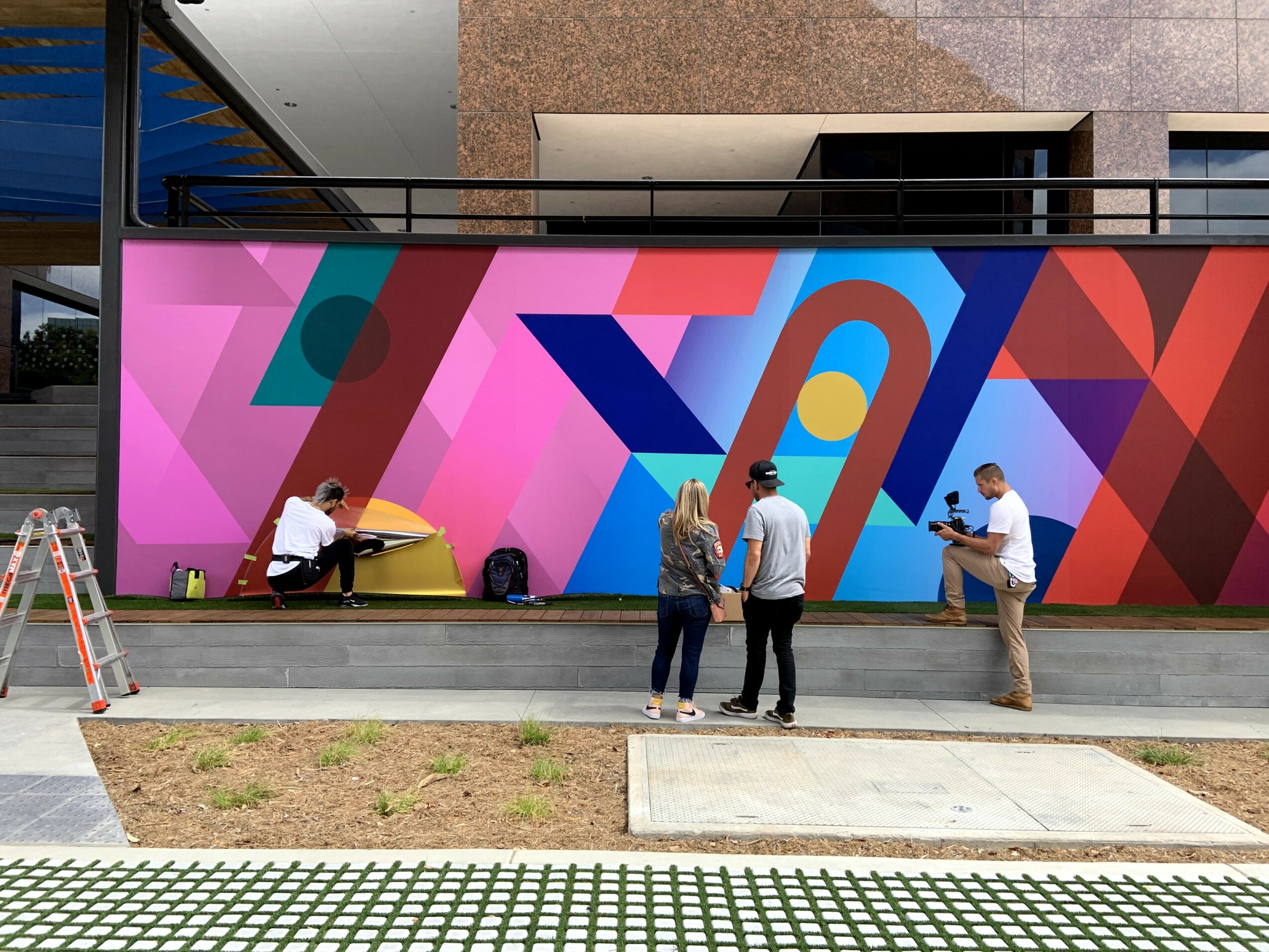 Four people near a colorful geometric mural, with one person sitting on a ledge and three standing and talking.