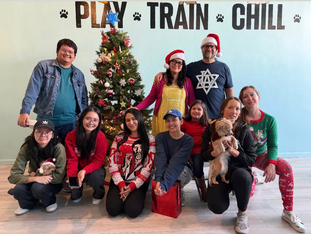 Group of nine people, some with dogs, in front of a decorated Christmas tree and holiday wall decor.