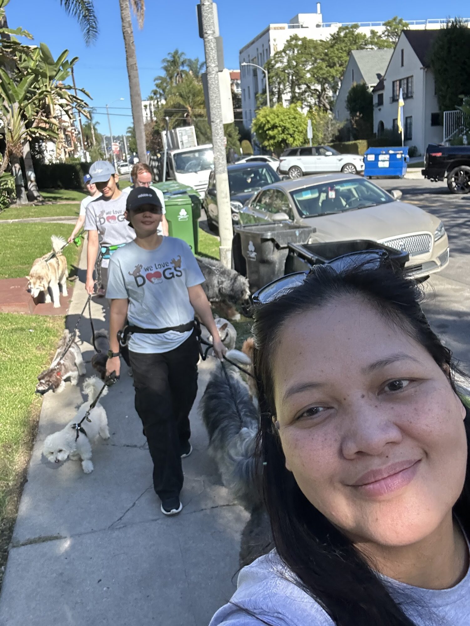 Woman taking selfie on sidewalk with people walking dogs, cars parked, and houses in background.