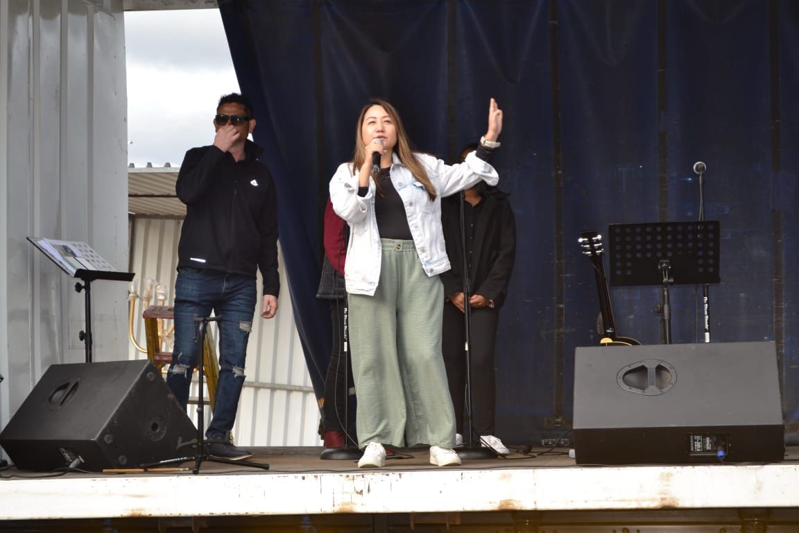 Three people on stage, one woman singing into a microphone, others standing behind her, stage setup with speakers and music stands.