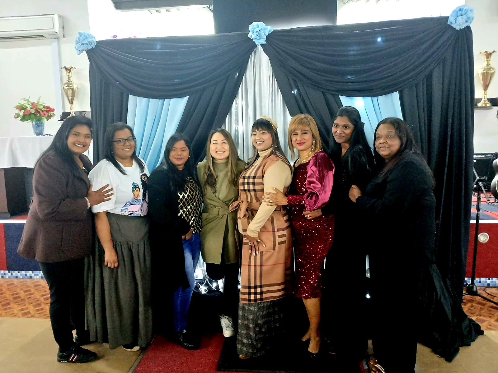 Group of eight women standing together indoors, with blue and black drapery backdrop, smiling for the photo.