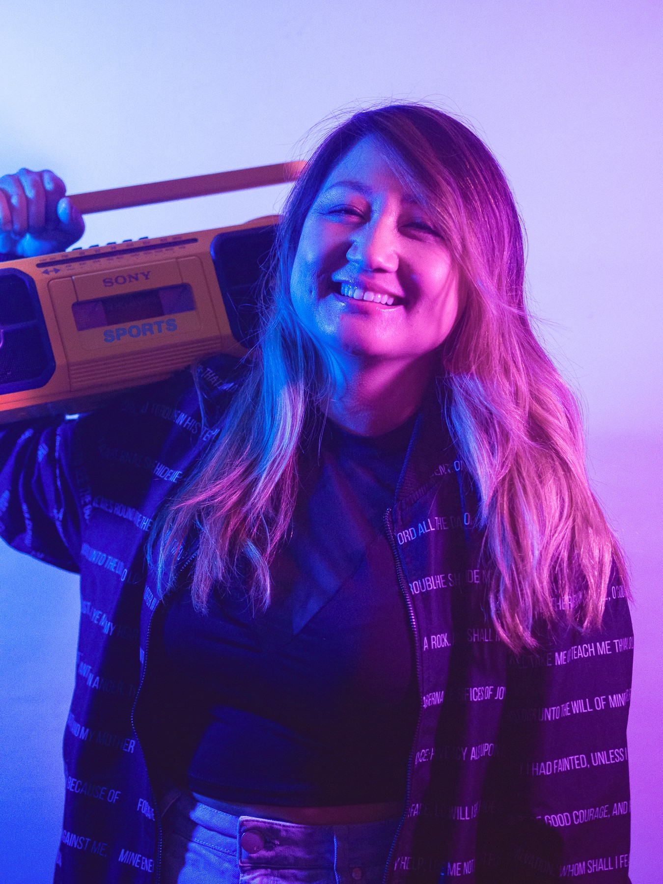 Smiling woman with long hair holding a boombox on her shoulder, colorful lighting background.