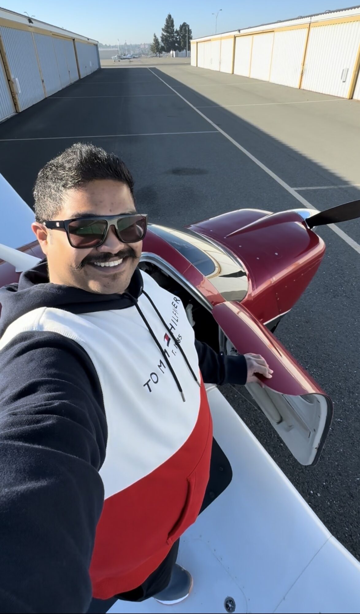 Person taking selfie next to small red aircraft on runway, wearing sunglasses and smiling.