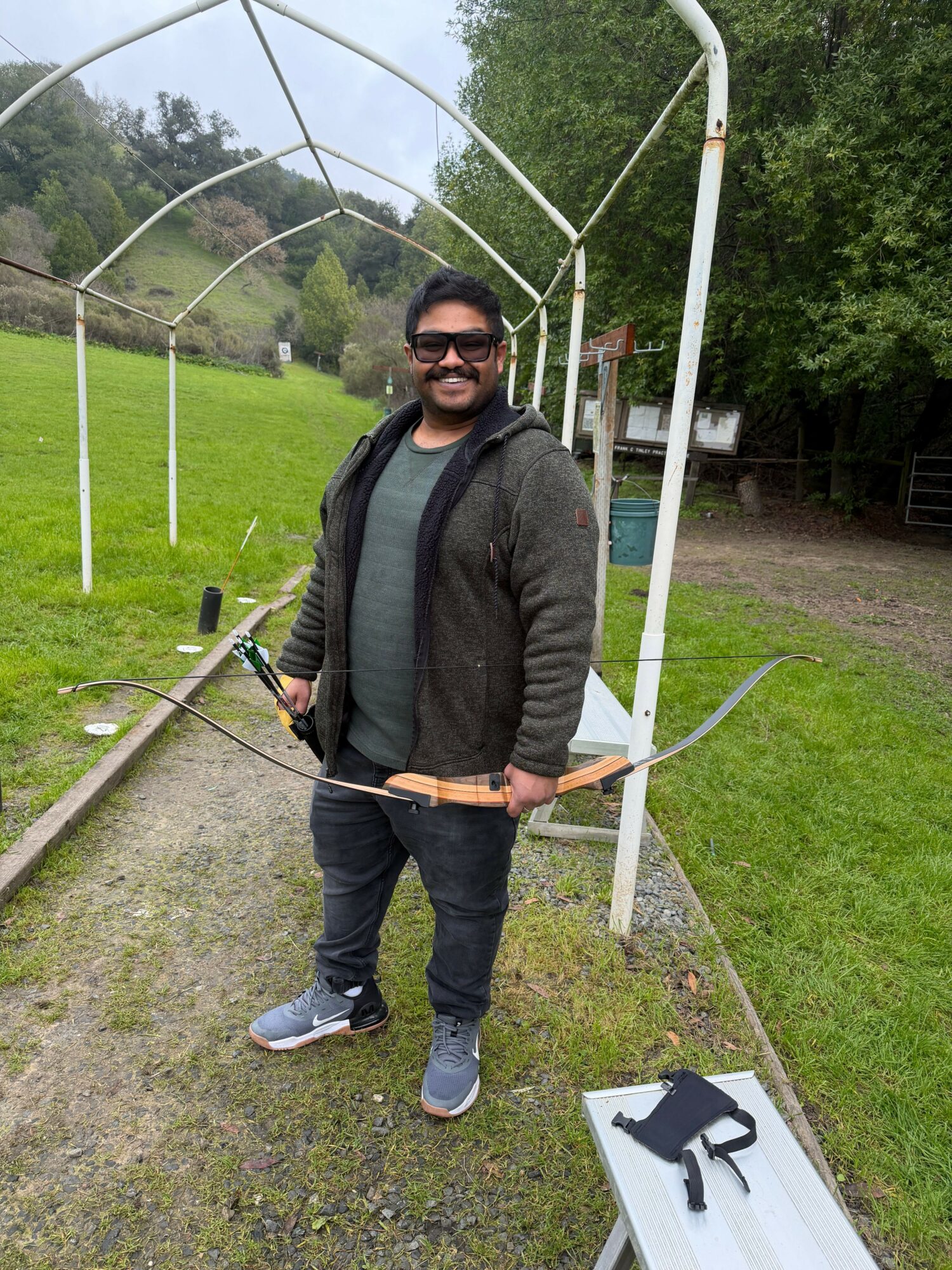 Man standing outdoors holding a bow, smiling, with a green field and trees in the background.