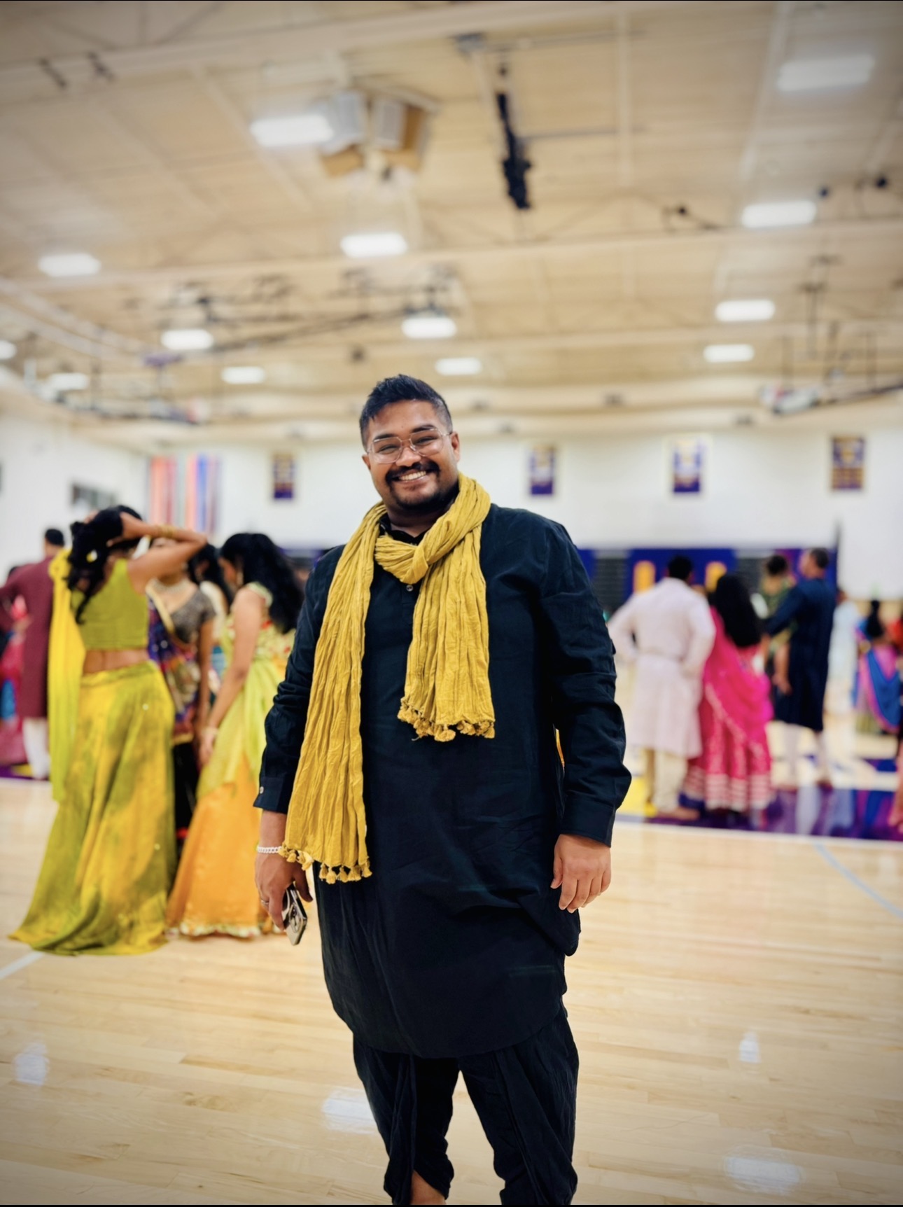 Smiling man in dark traditional attire with yellow scarf in a gymnasium with people in colorful clothing.