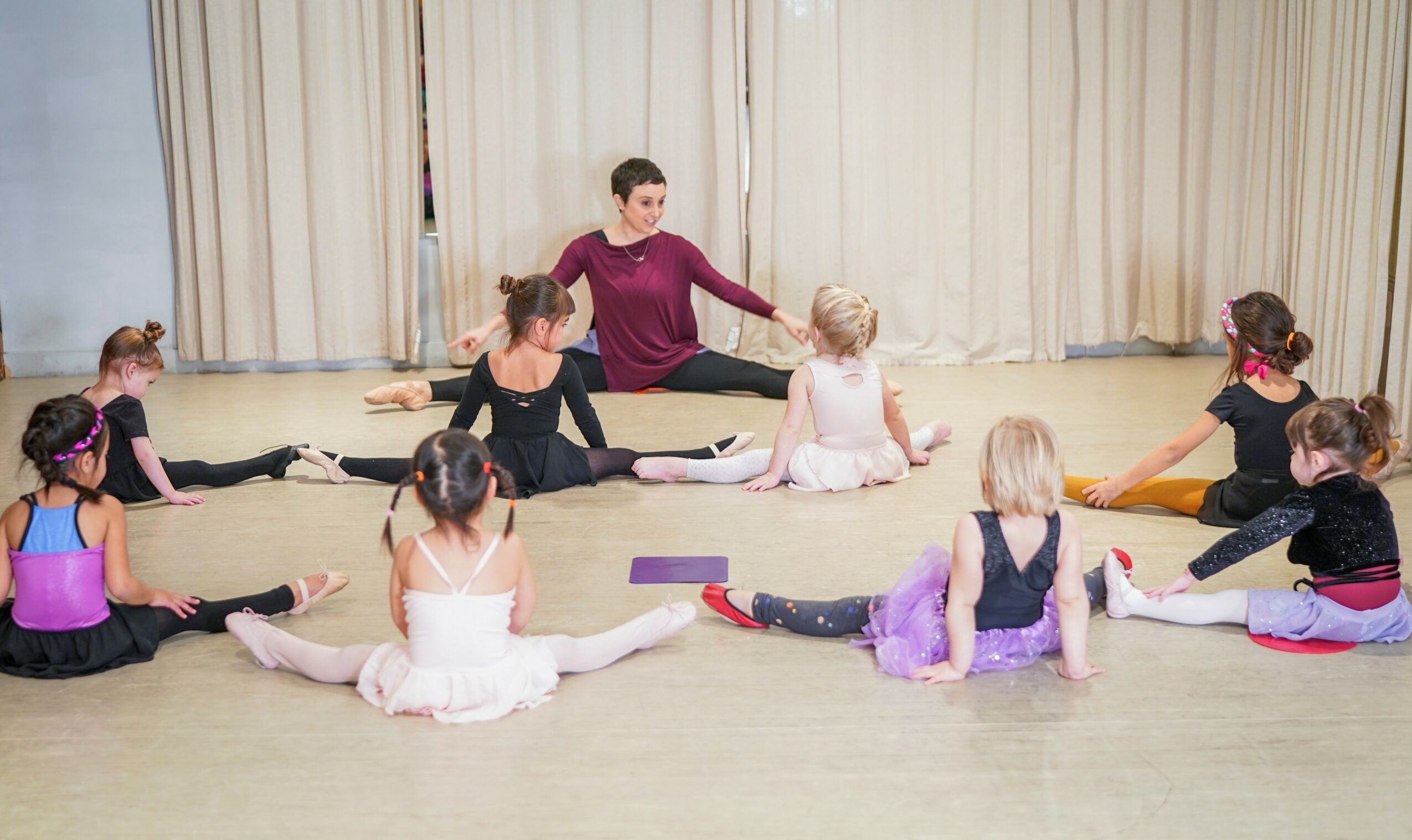 Instructor and children in a dance class sitting in a circle on the floor, stretching legs apart.