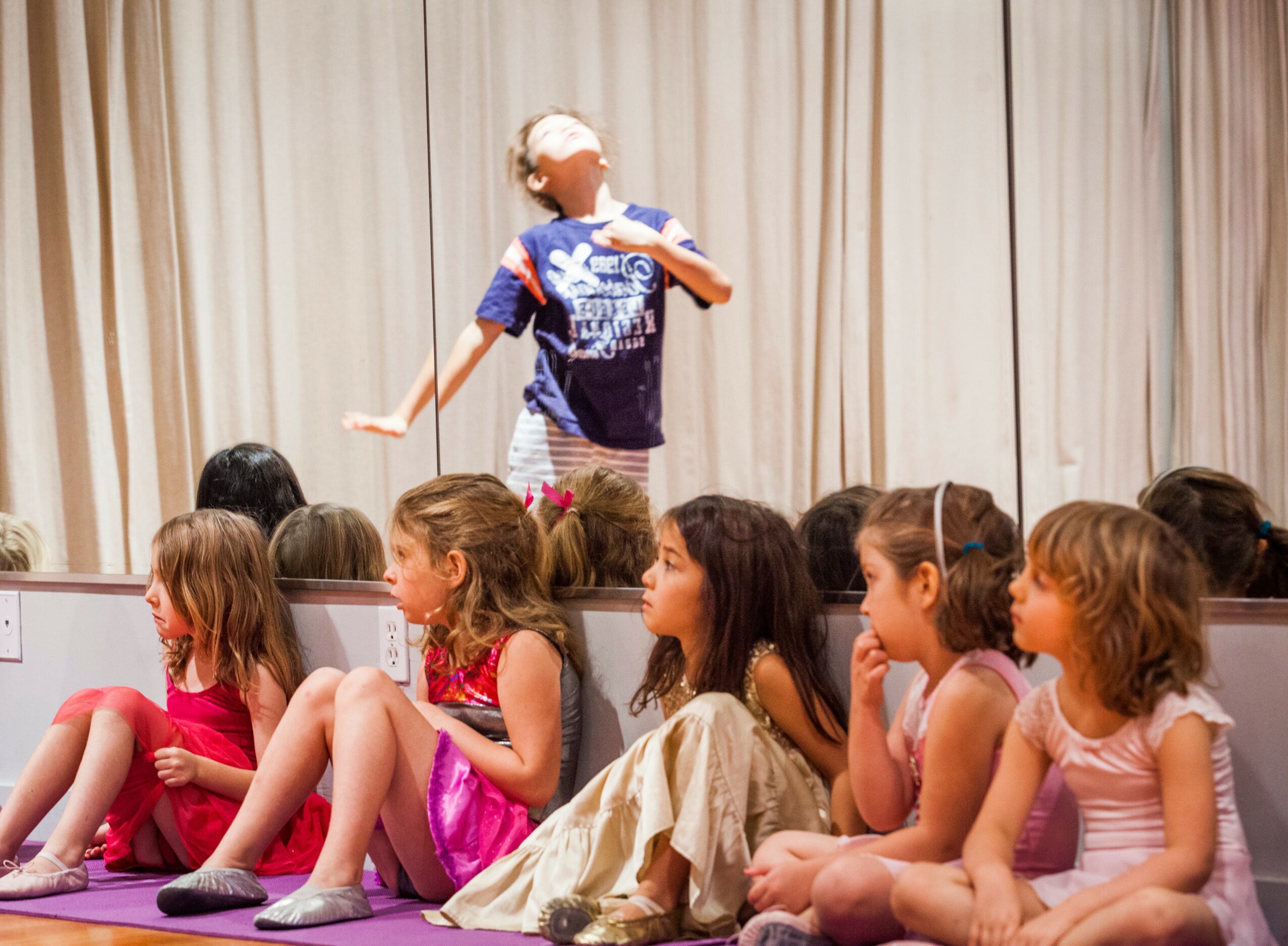 Child standing on stage with a microphone, other children sitting on the floor watching, curtain backdrop.