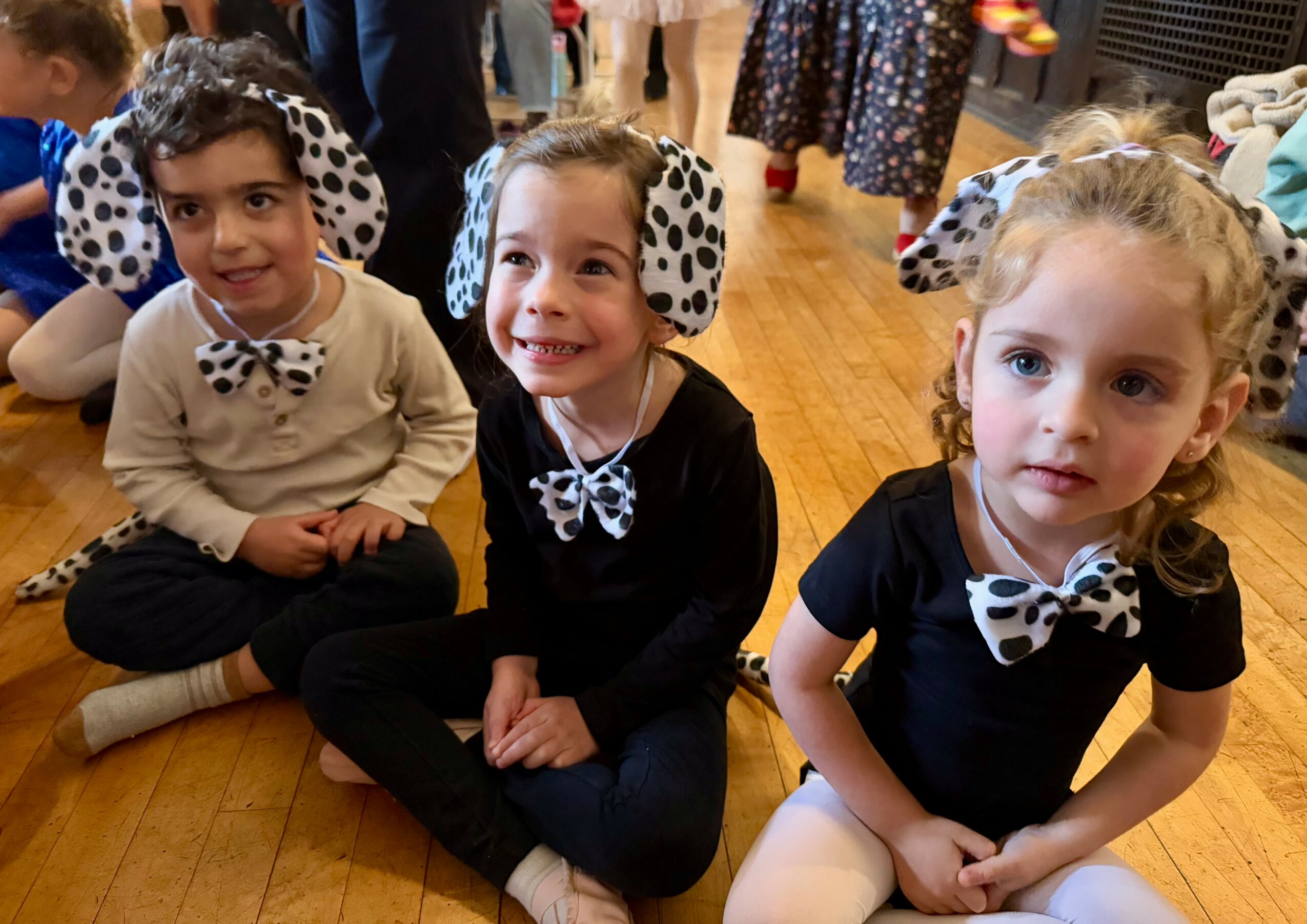 Three children sitting on a wooden floor, wearing black and white polka dot headbands and bow ties, smiling.