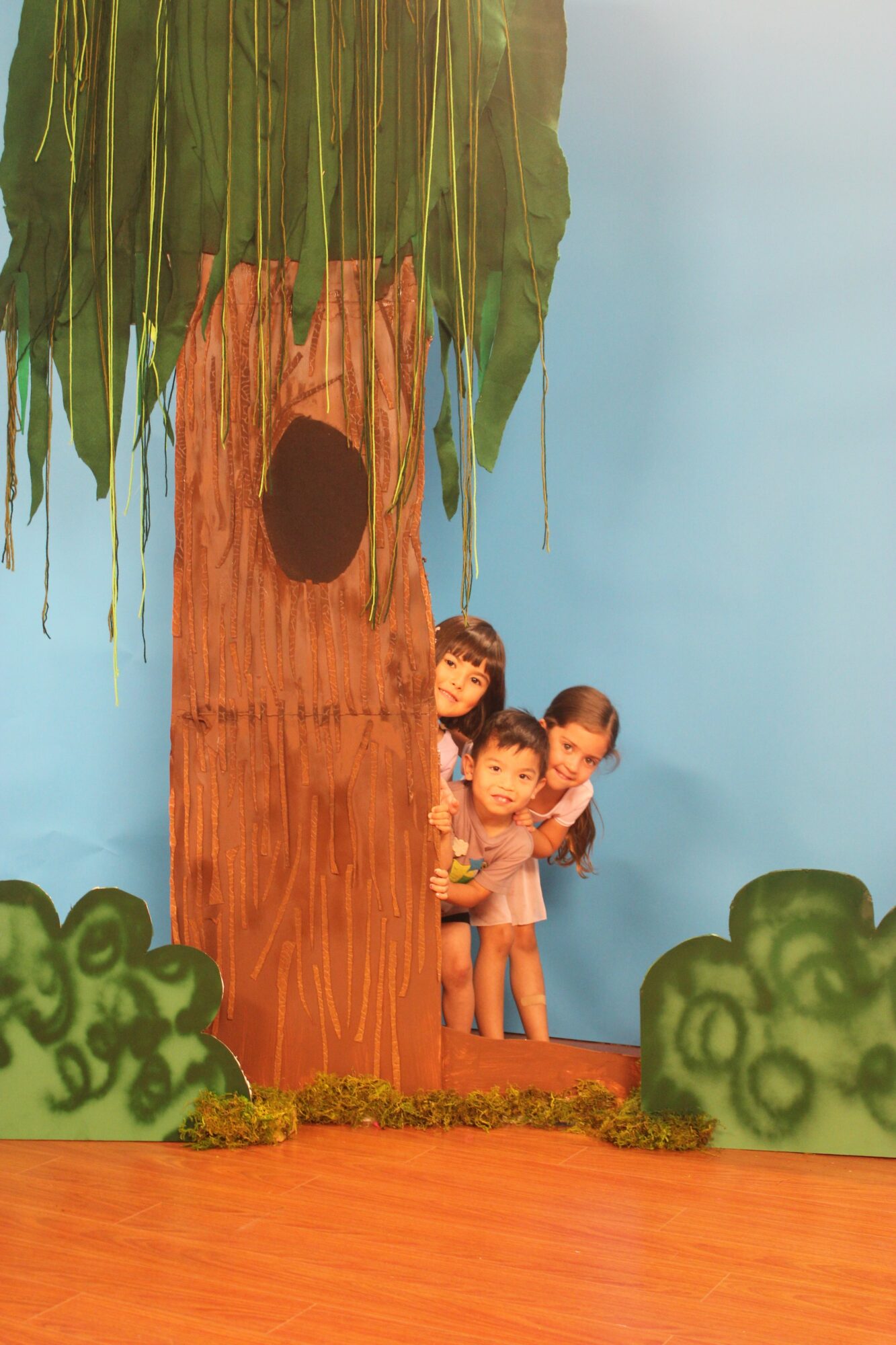 Three children peek from behind a large tree with green leaves, on a colorful stage with bushes and a blue background.