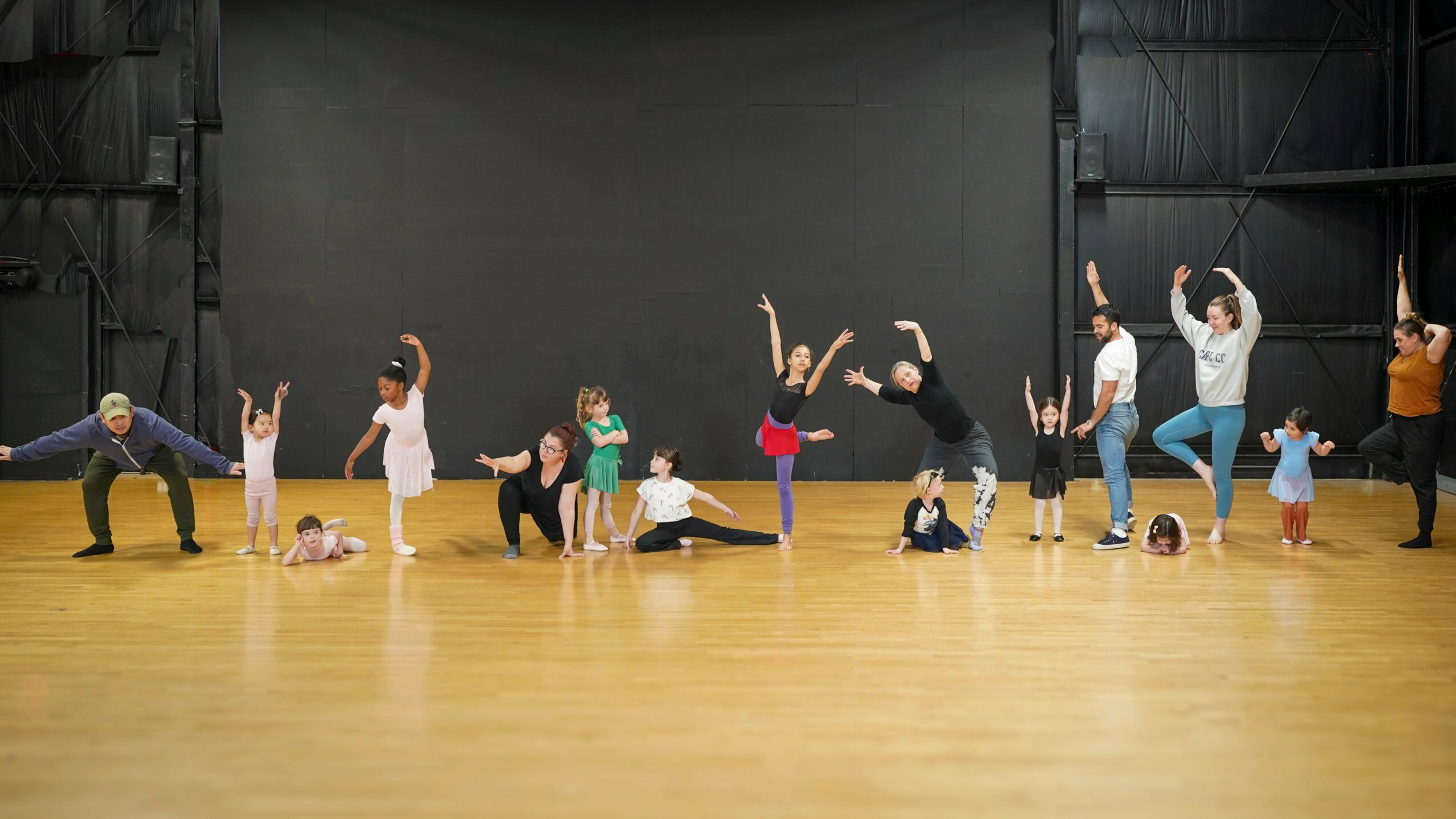 Group of children and adults in a dance studio, some stretching and practicing dance moves on a wooden floor.