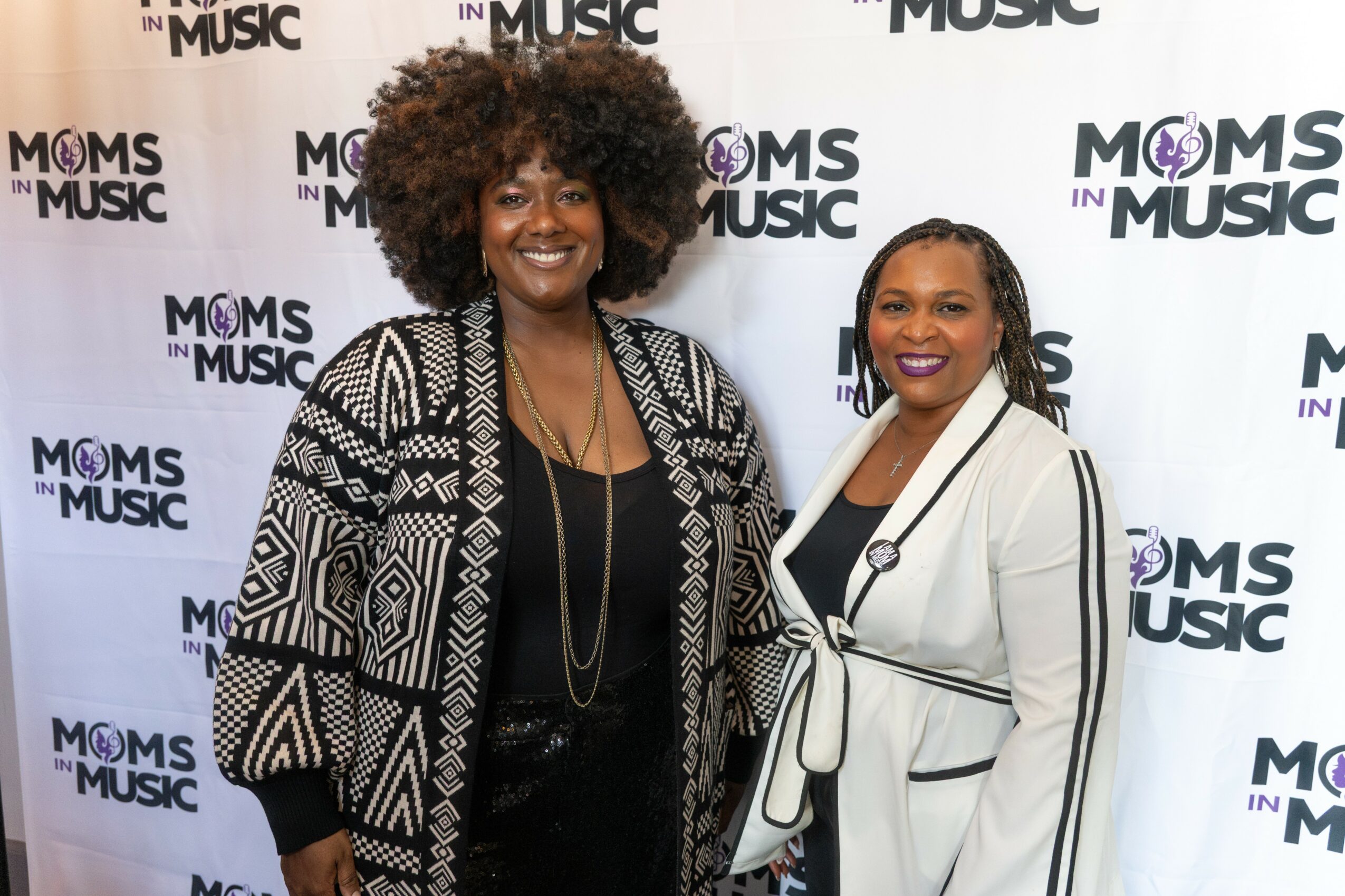 Two women standing in front of a backdrop with 'Moms in Music' logo, smiling, wearing black and white outfits.