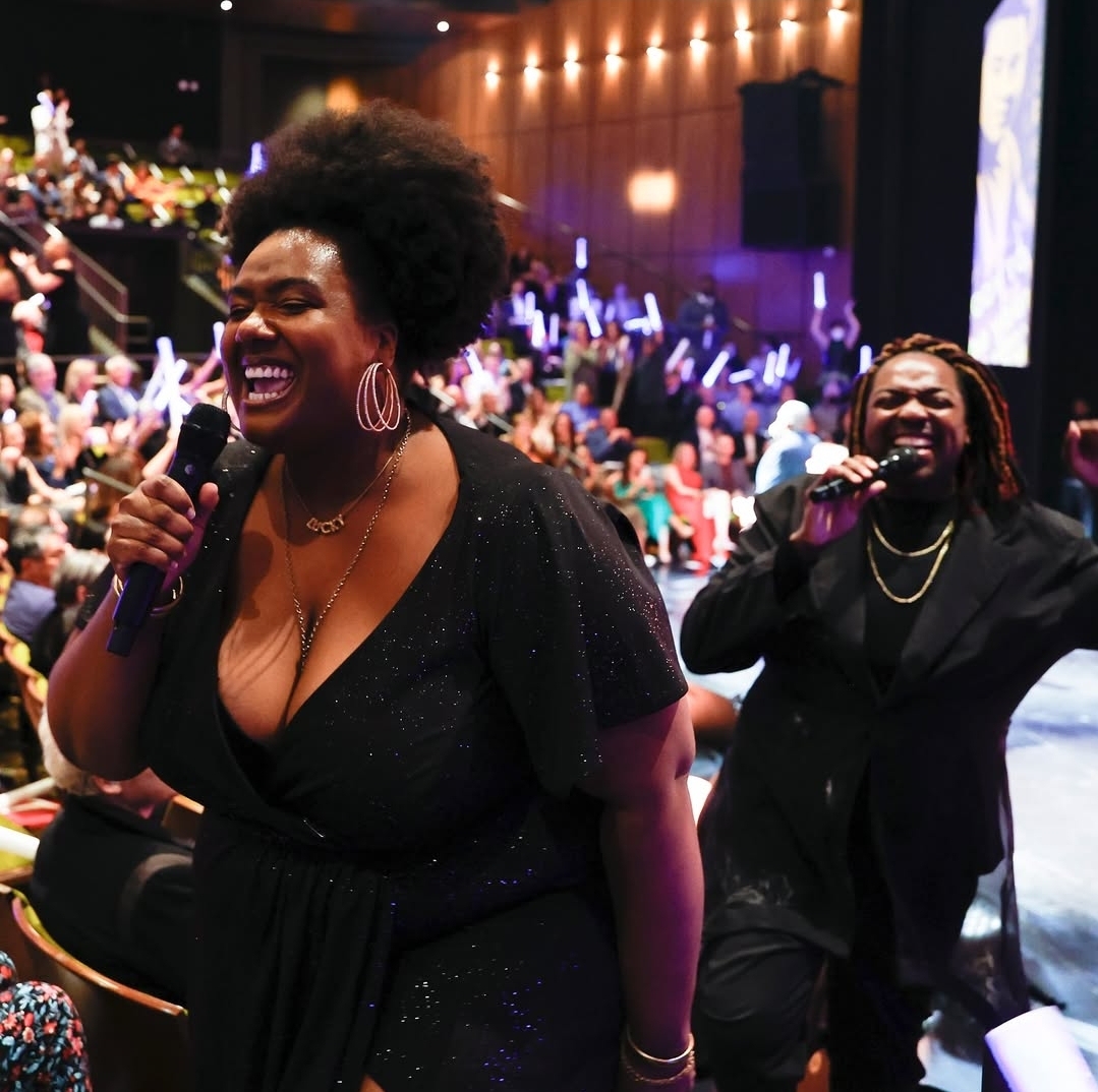 Two women singing and smiling on stage with an audience in the background.