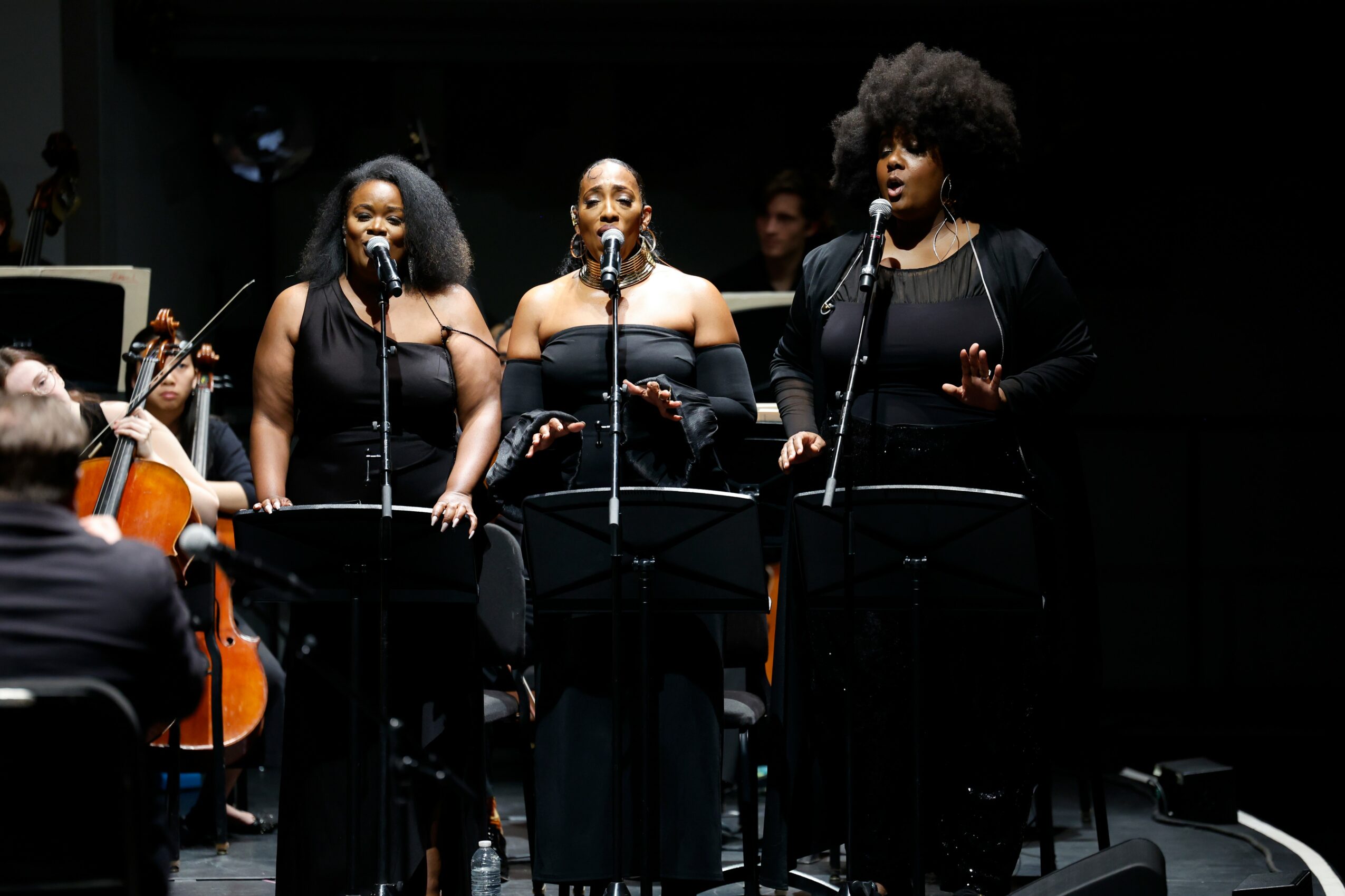 Three women sing into microphones on stage with a band and orchestra in the background.