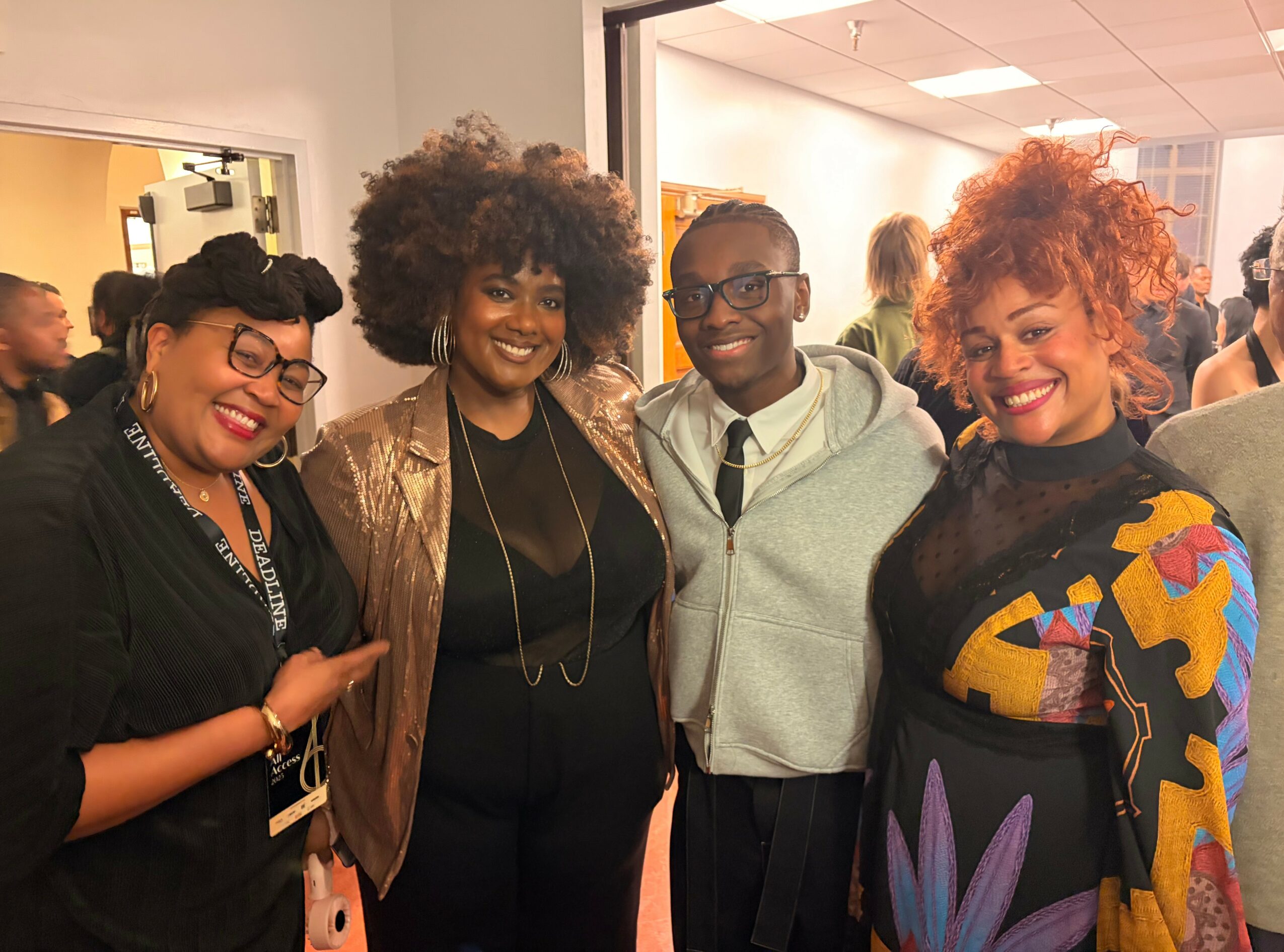 Four people smiling and posing together indoors, diverse hairstyles and colorful clothing, background with other people.
