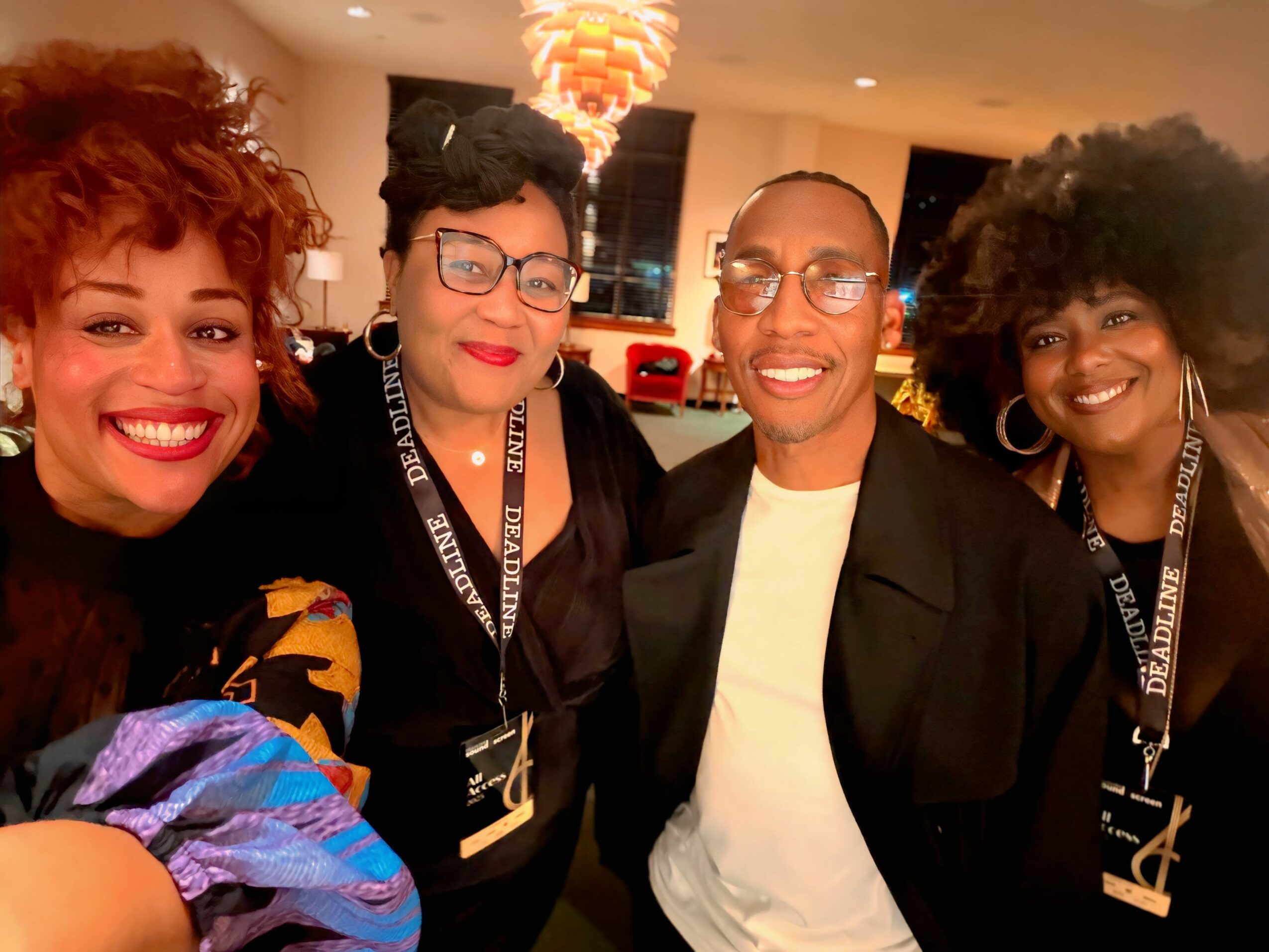Four people smiling and posing together indoors, three women and one man, with warm lighting and modern decor.
