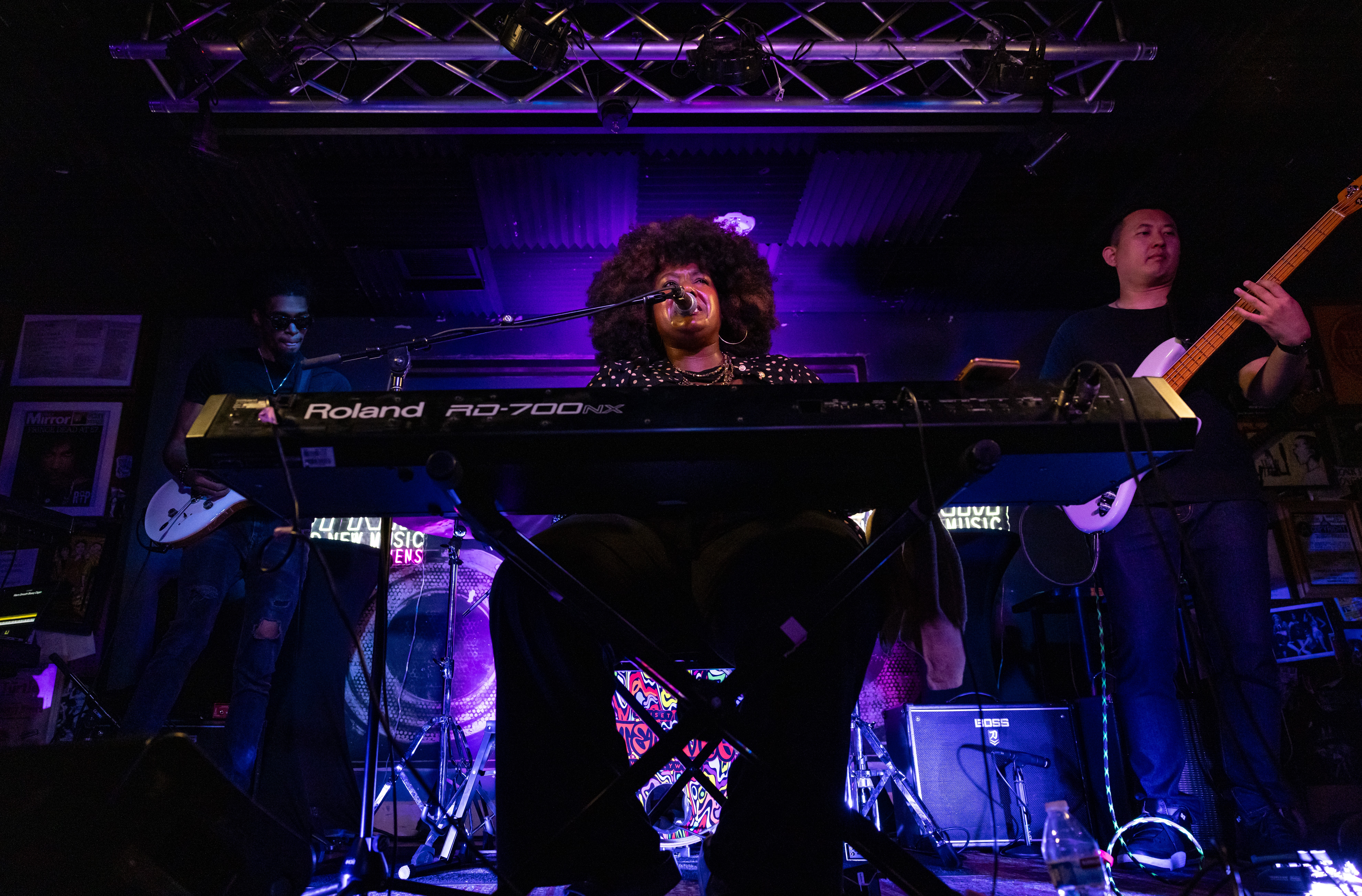 Musician with curly hair playing keyboard on stage with band members in background.