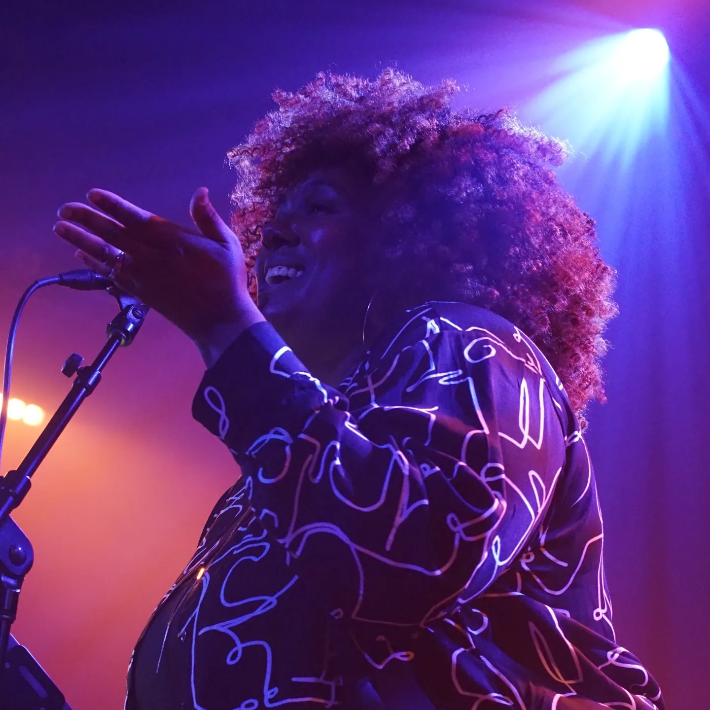 Person with curly hair singing into a microphone on stage with colorful lighting.