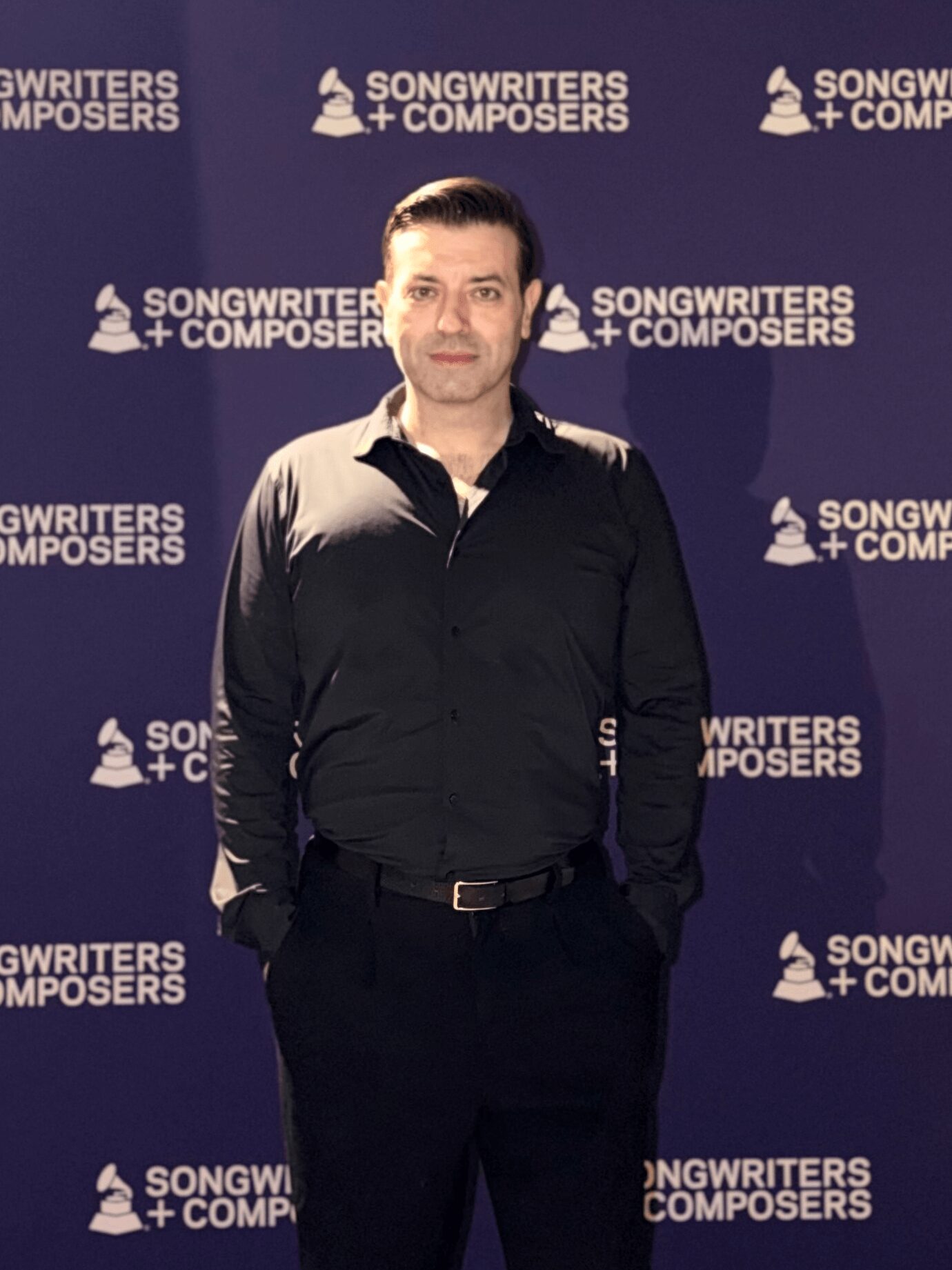 Man in black shirt standing in front of a backdrop with 'Songwriters + Composers' logo.