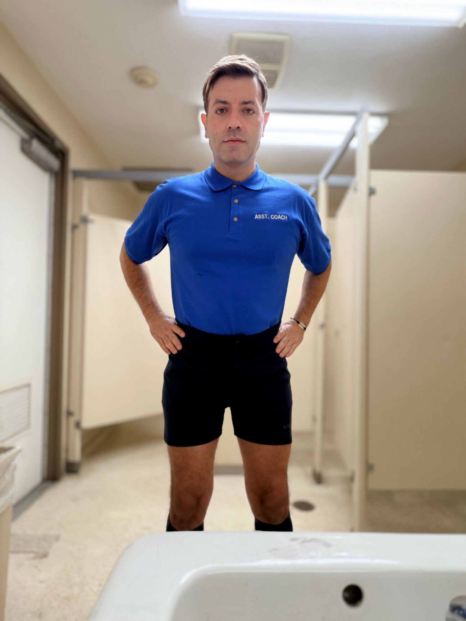 Man in blue polo shirt and black shorts standing in a room with beige walls and white ceiling, hands on hips.