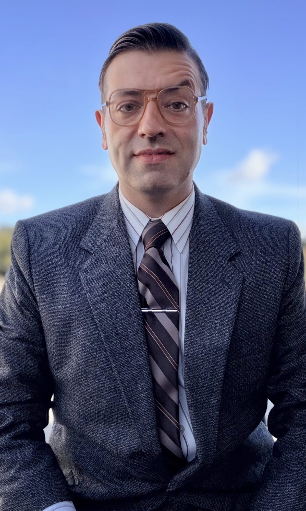 Man in suit and tie outdoors with blue sky and clouds behind him.
