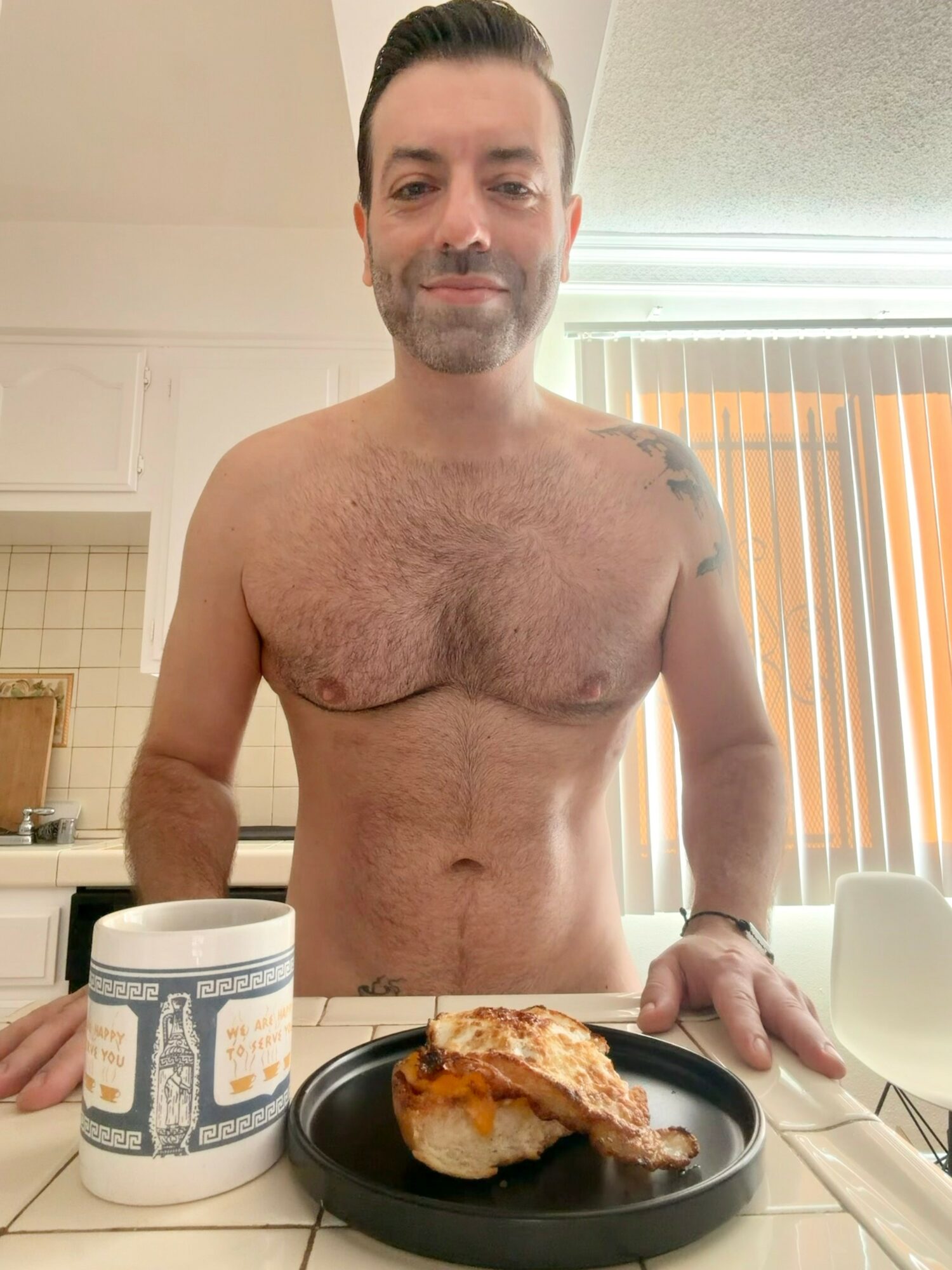 Man with short dark hair and beard, shirtless, standing in kitchen with food on table, smiling.