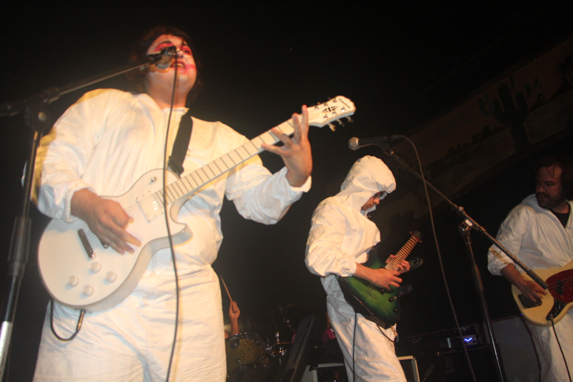 Three musicians performing on stage, two playing guitars and one singing, dressed in white outfits, with dark background.