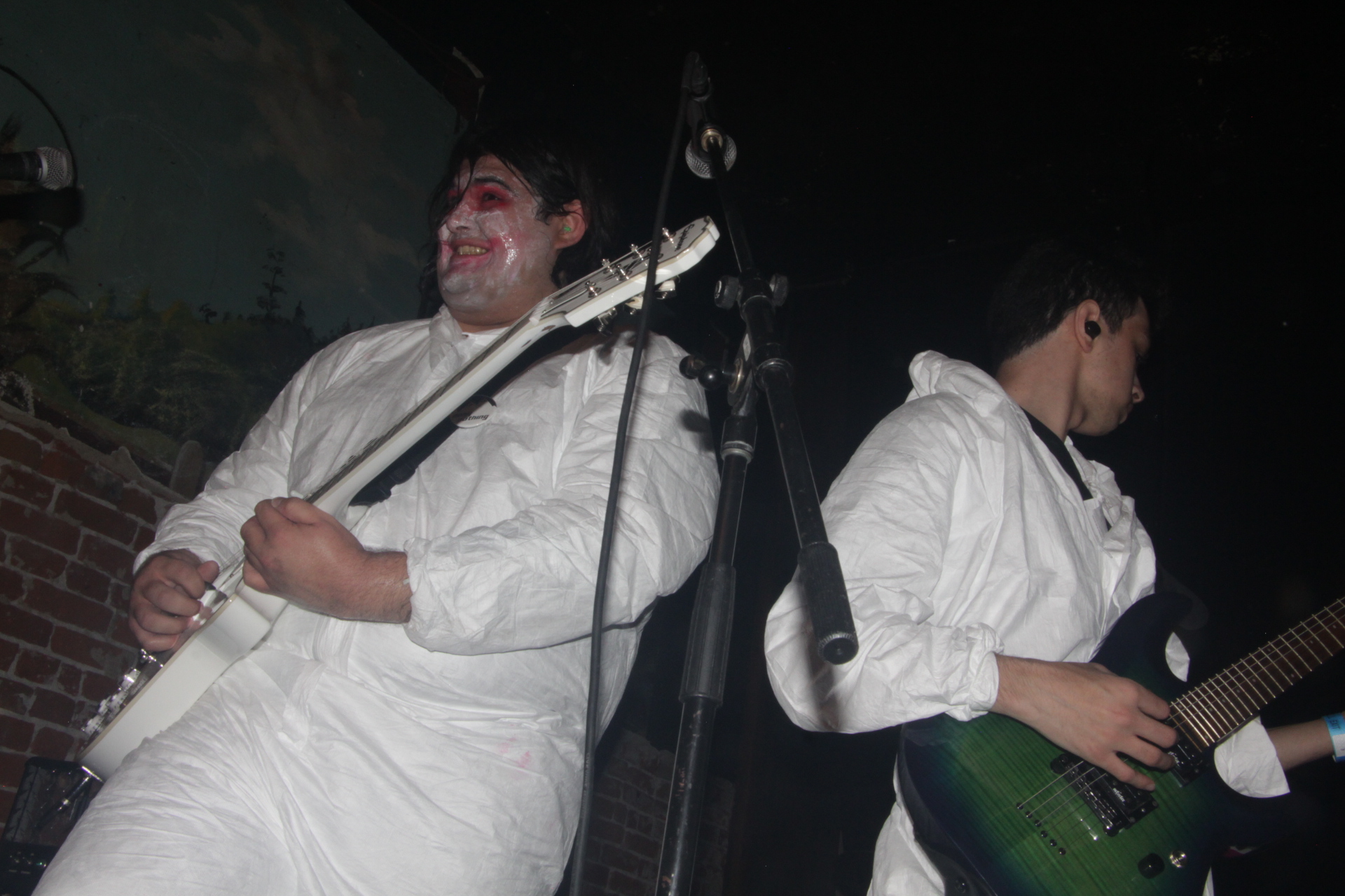 Two people in white costumes on stage, one smiling and holding a guitar, the other looking down with a guitar.