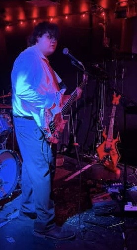 Person with curly hair holding an electric guitar on stage with microphone and other guitars nearby.