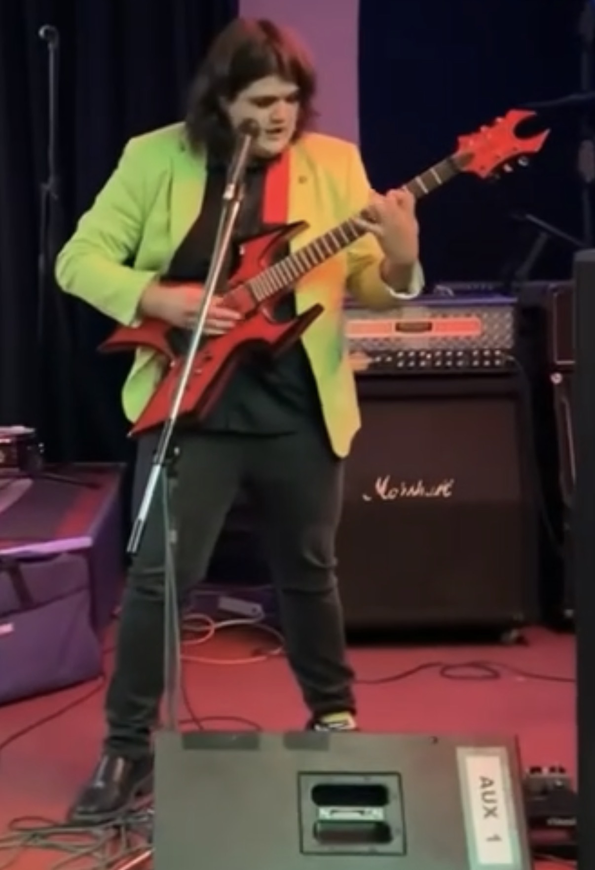 Person playing red electric guitar on stage with amplifier and microphone, wearing a bright green jacket and black pants.
