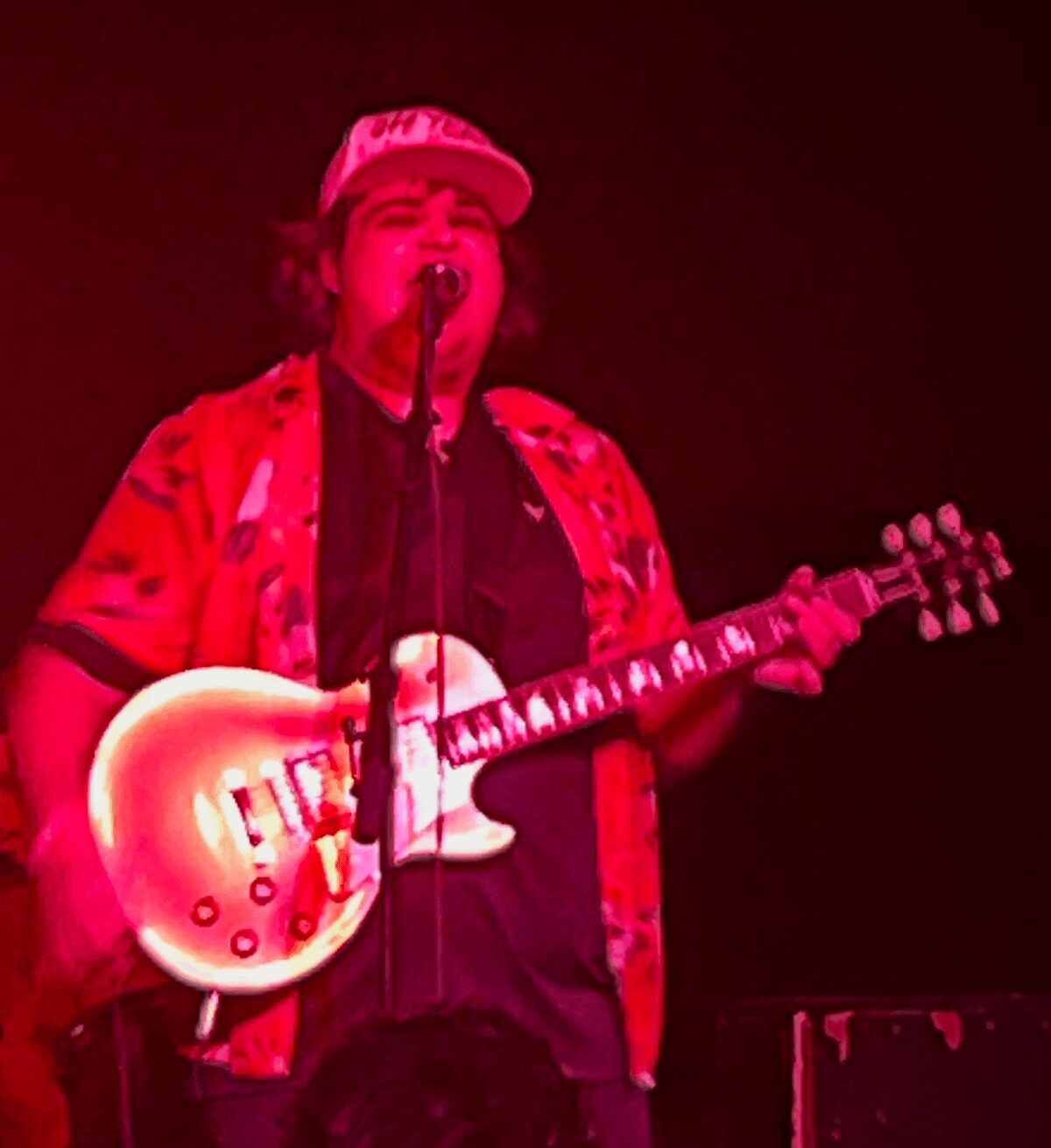 Person singing into microphone while playing guitar, wearing a cap and colorful shirt, on stage with dark background.