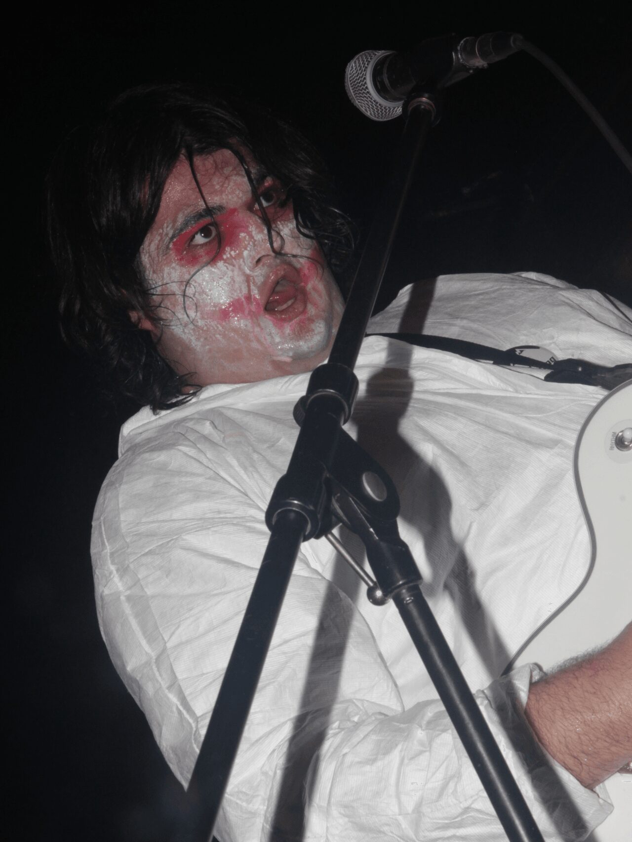 Person with face paint or makeup, wearing a white shirt, speaking into a microphone on stage.