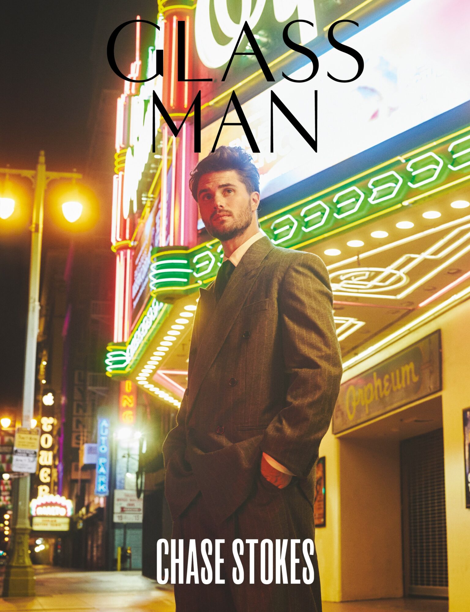Man in a suit standing on a city street at night with neon lights and signs behind him.