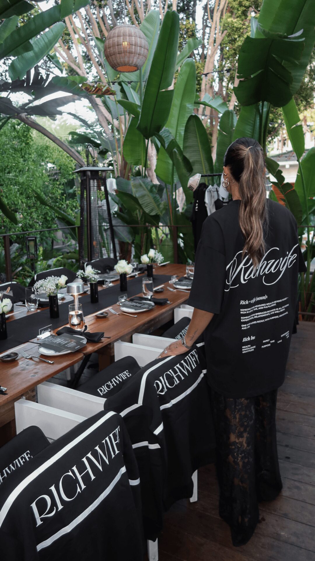Person with long hair wearing a black oversized shirt in a tropical outdoor setting with tables and chairs.