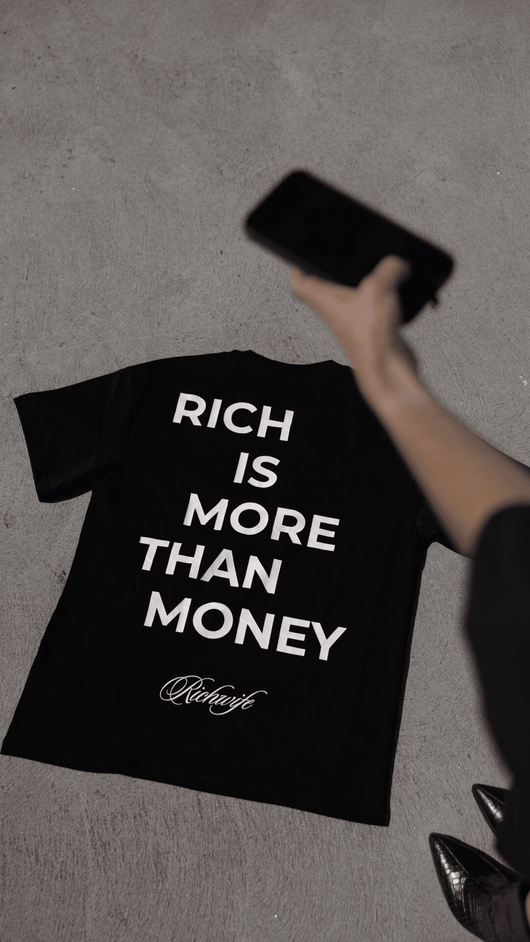 Black T-shirt with white text reading 'Rich is more than money' and a logo at the bottom, laid on a gray surface.
