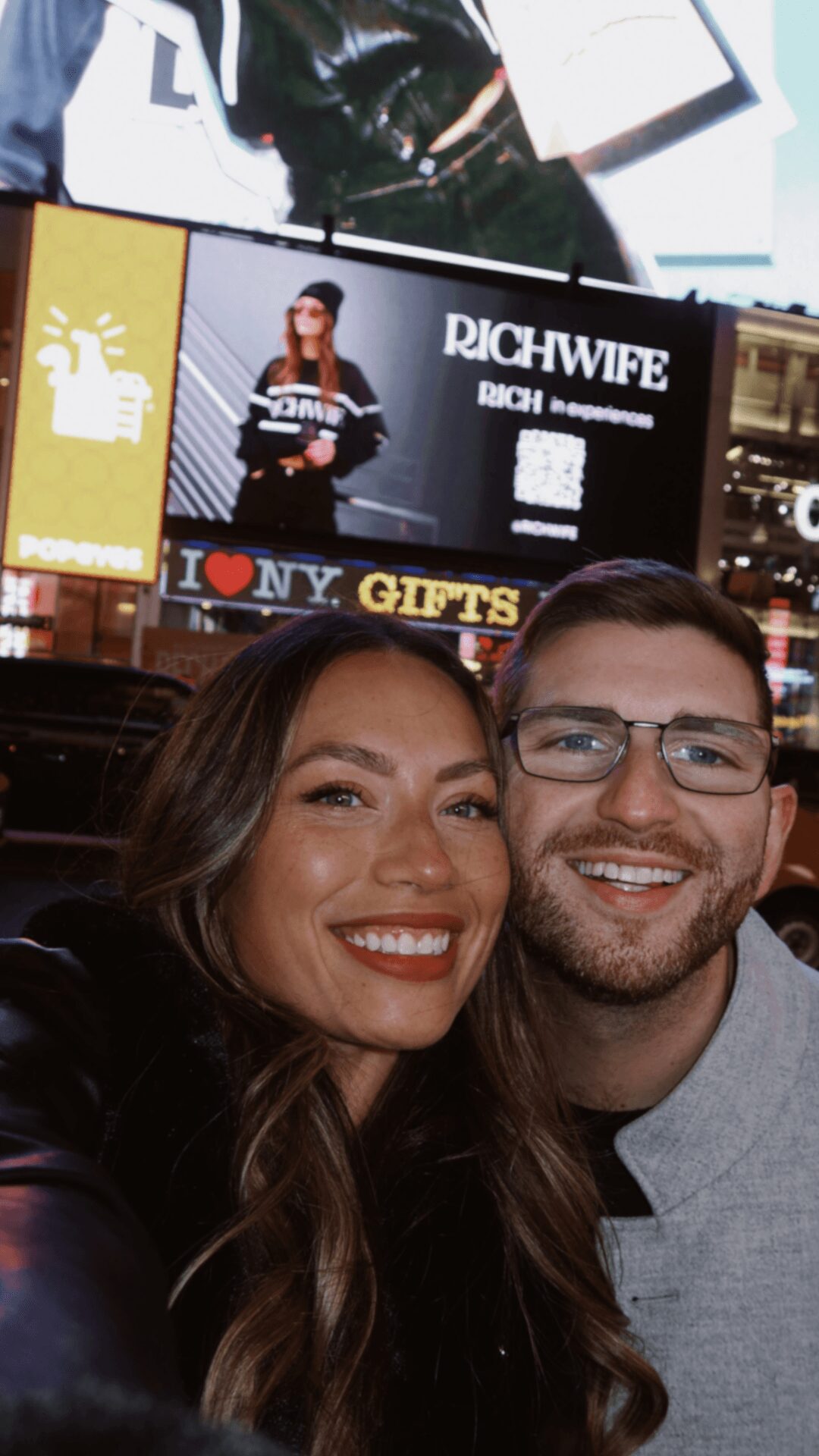 Two smiling people taking a selfie in a busy indoor setting with screens and signs in the background.
