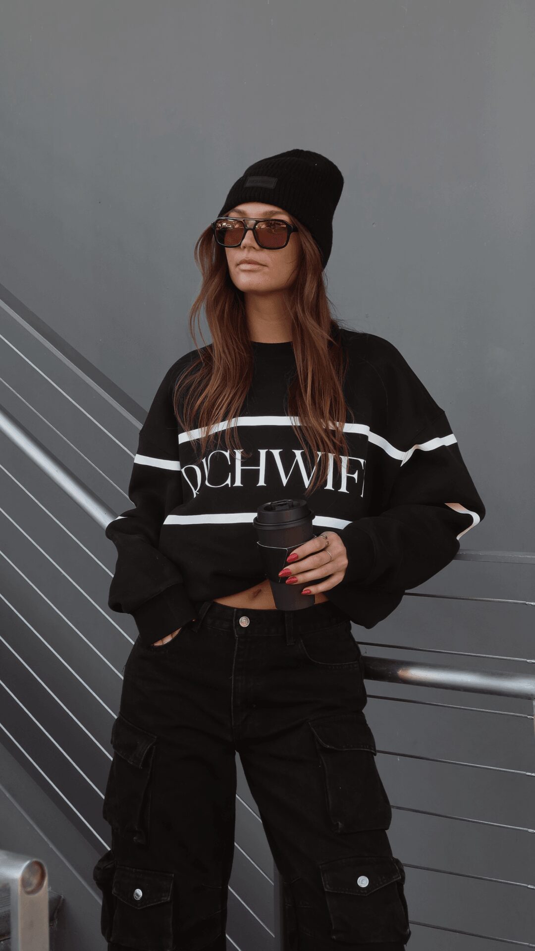 Woman wearing sunglasses, beanie, black sweatshirt with white stripes, and black pants, holding a cup, standing outdoors.