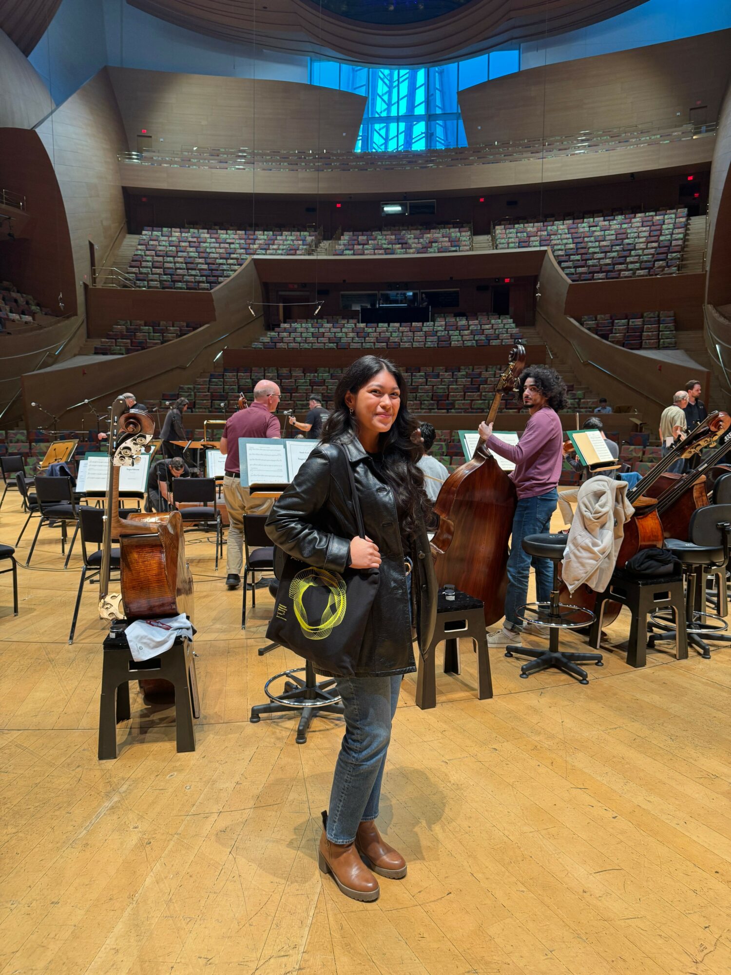 Woman standing in a large auditorium with chairs and people, stage and balcony in background.