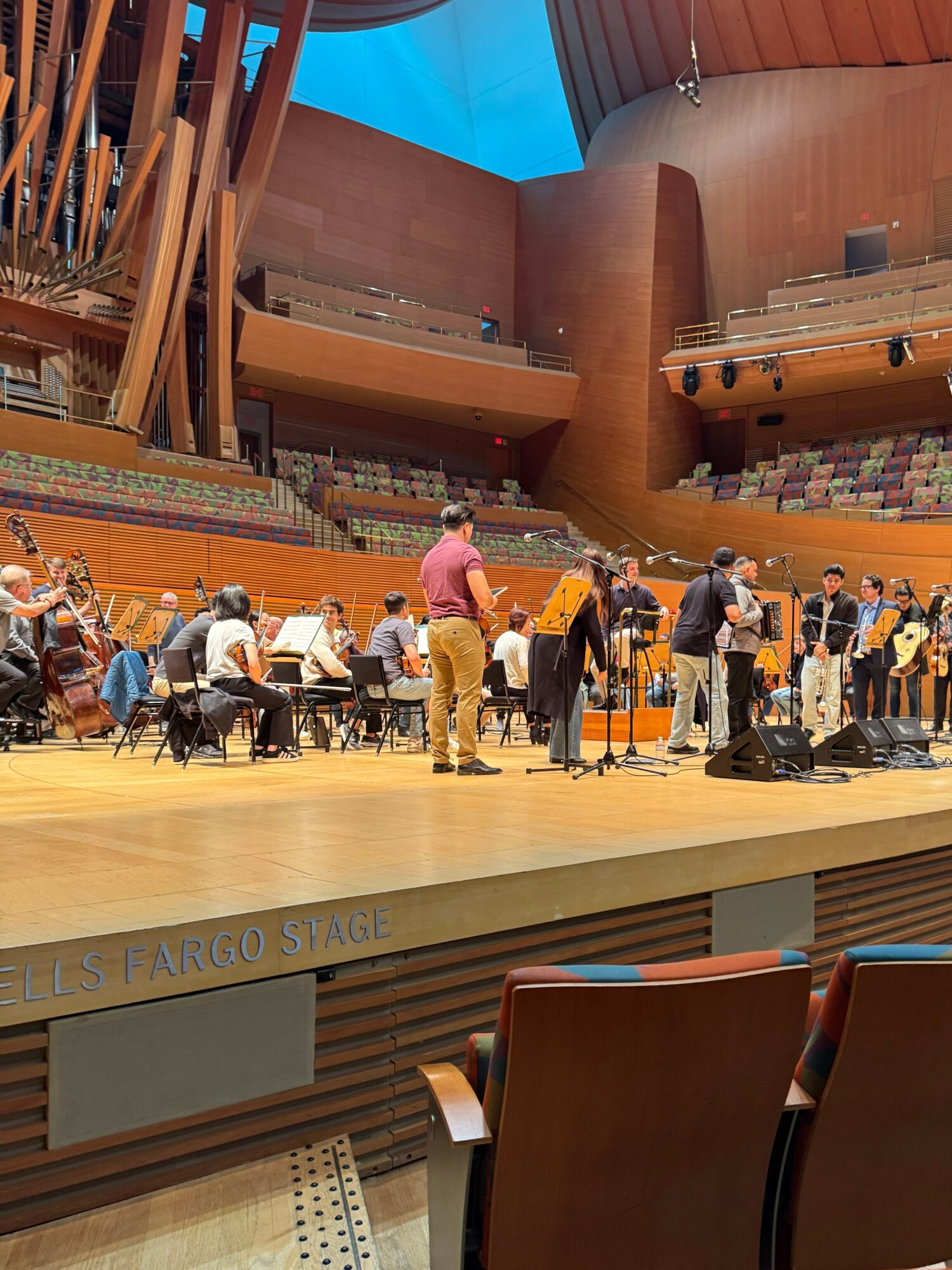 Orchestra performs on stage with musicians and chairs in a large concert hall with wooden architecture.
