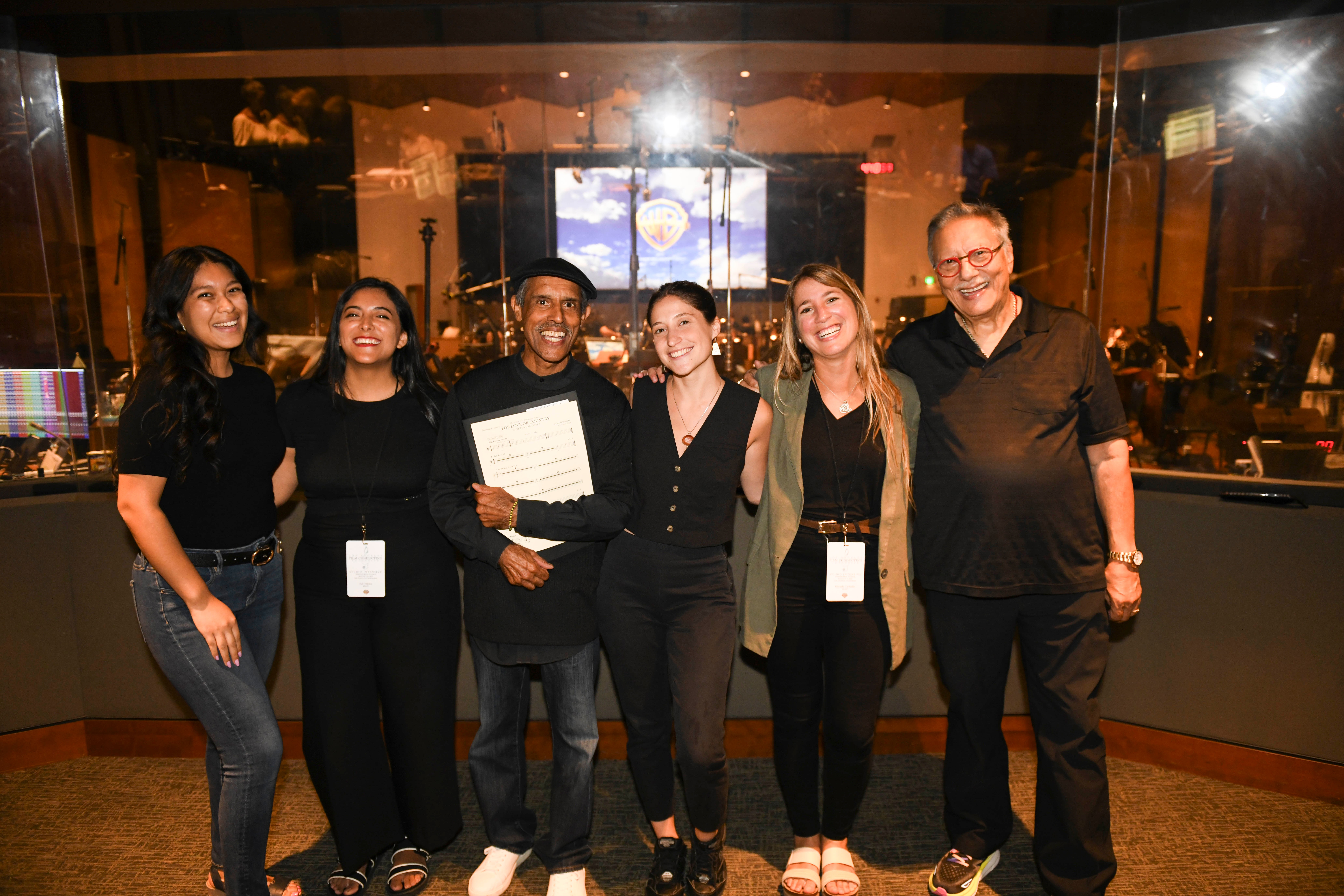 Six people standing together indoors, smiling, with city lights visible through windows behind them.