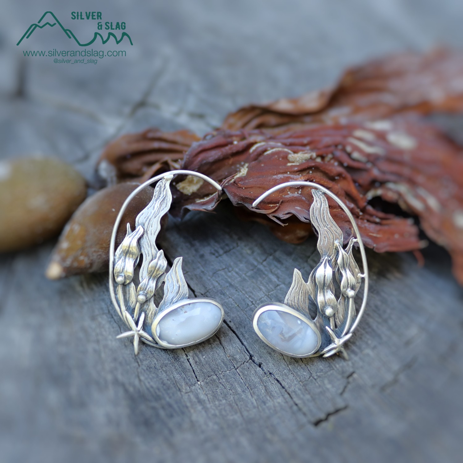 Silver bracelet with leaf design and oval stones, placed on a wooden surface with dried leaves and stones nearby.