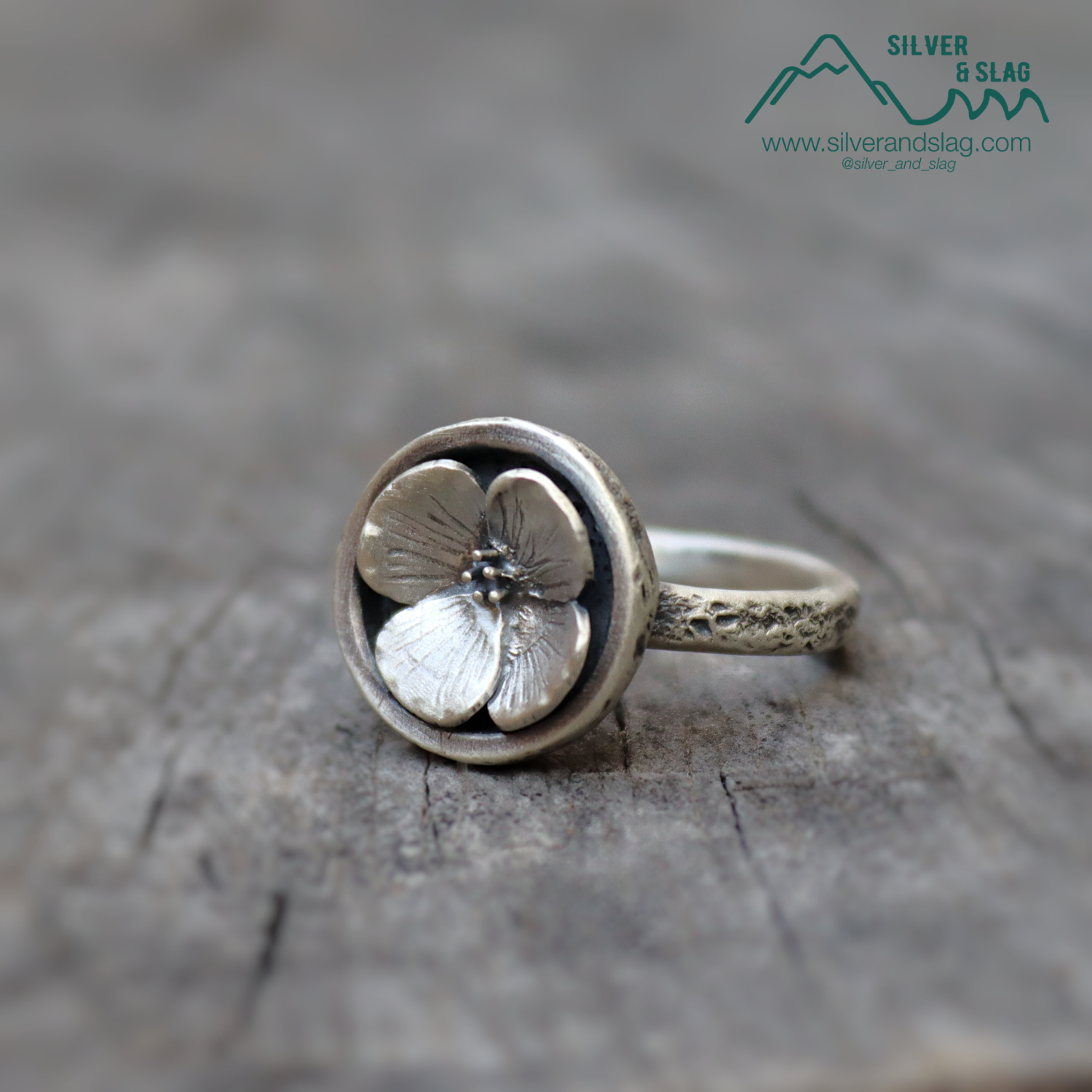 Silver ring with a flower design featuring four petals, placed on a textured surface.