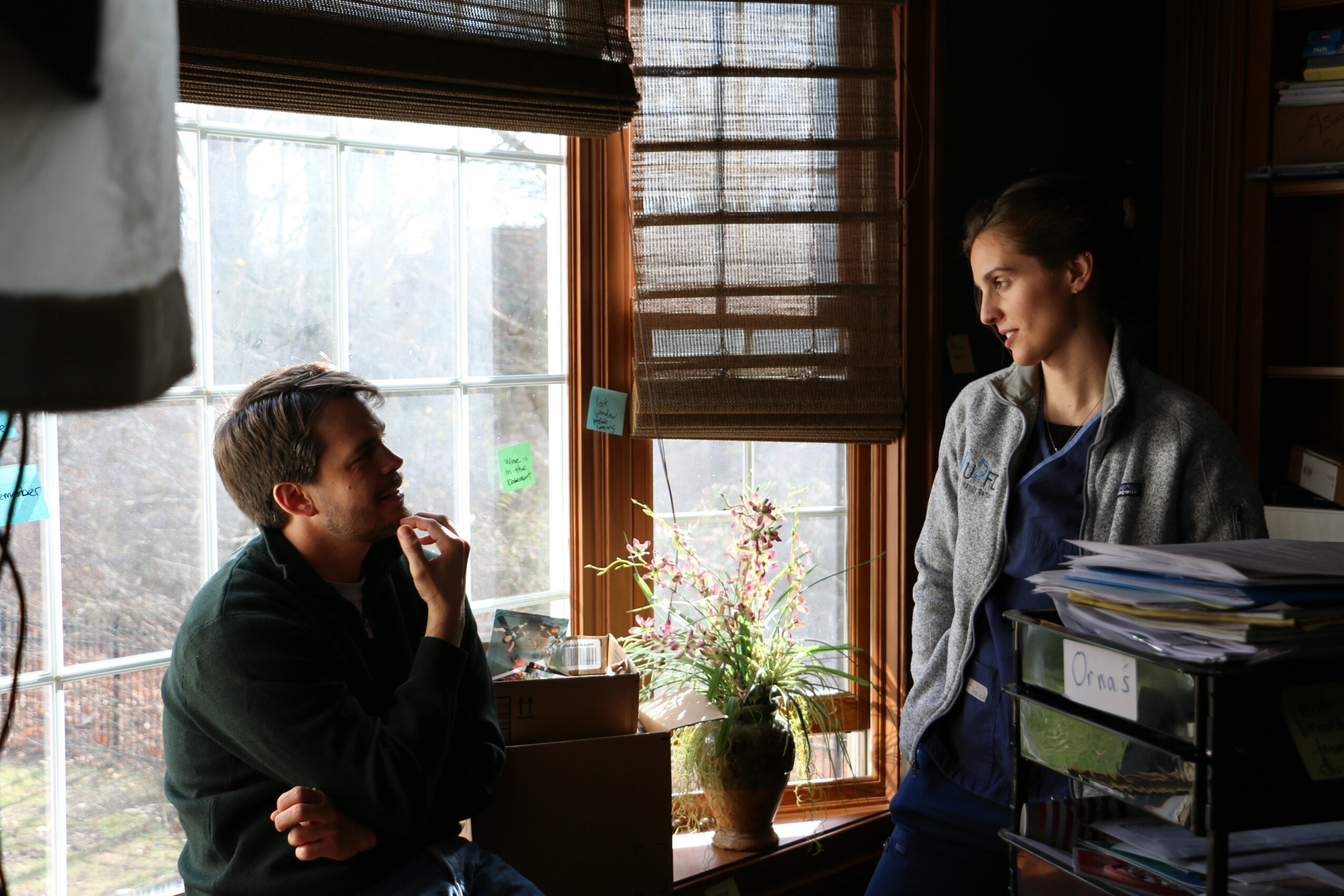 Two people talking indoors near a window with a plant and sunlight, one sitting and one standing with a stack of papers.