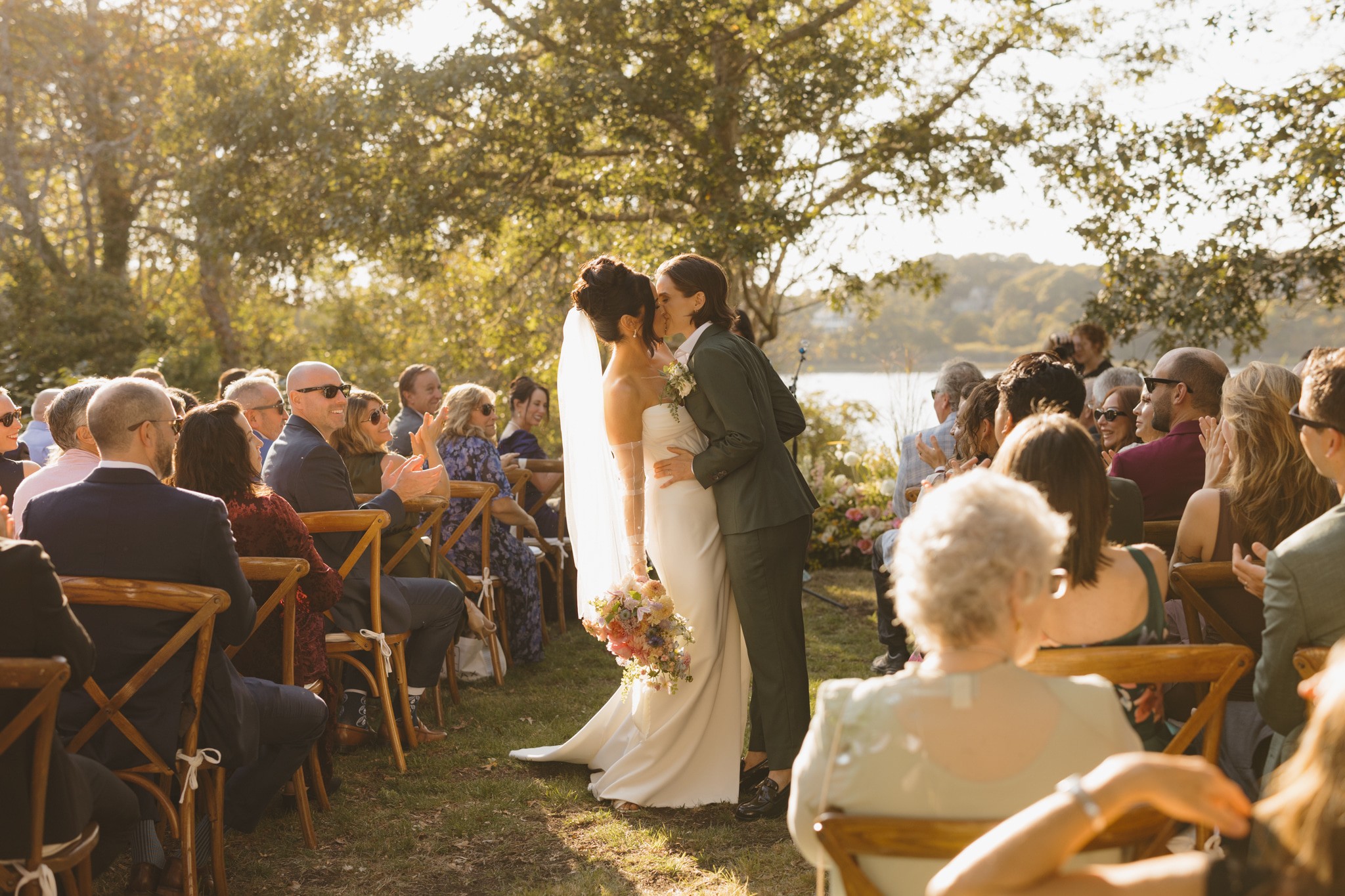 Bride and groom kiss at outdoor wedding ceremony with seated guests and trees in background.