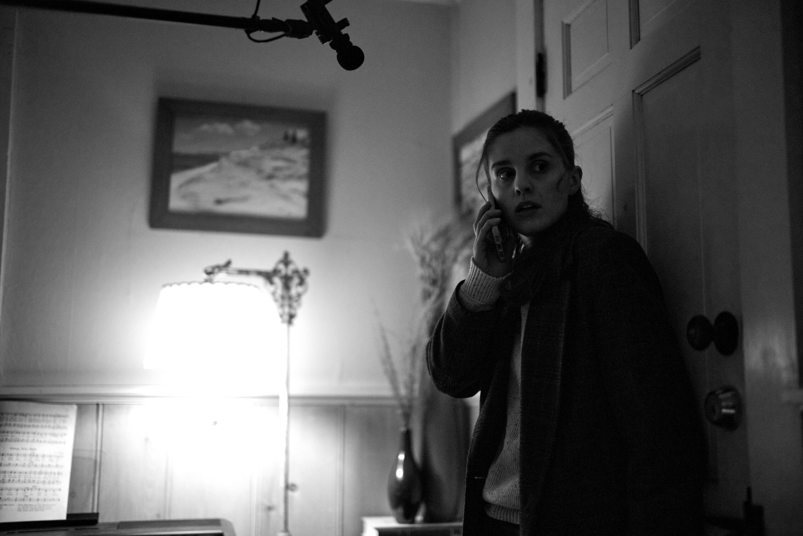 Girl standing near door, talking on phone, in a room with a lamp and framed picture on wall.