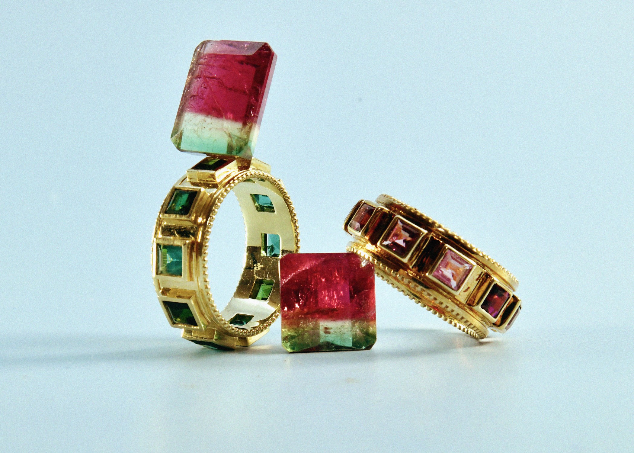 Three gold rings with embedded gemstones and two watermelon slices, one on each ring, on a light blue background.