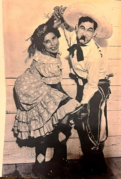 A man and woman in vintage costumes dance together, holding hands, smiling, with a painted backdrop behind them.