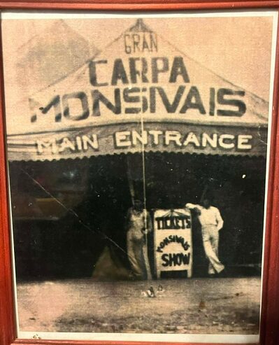 Two people stand outside a tent with a sign that reads 'Main Entrance' and 'Tents.' The tent has a sign for a show.