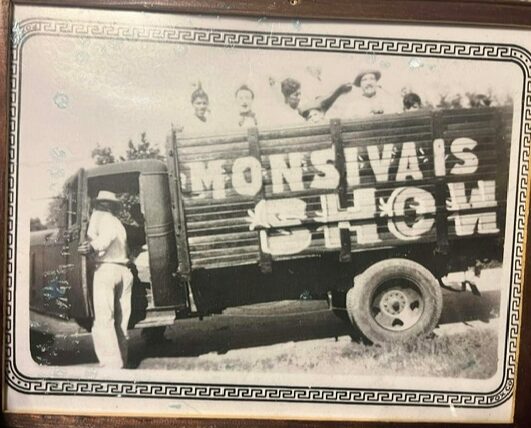 People on a truck with large painted letters, some standing and one leaning against the truck, outdoors.