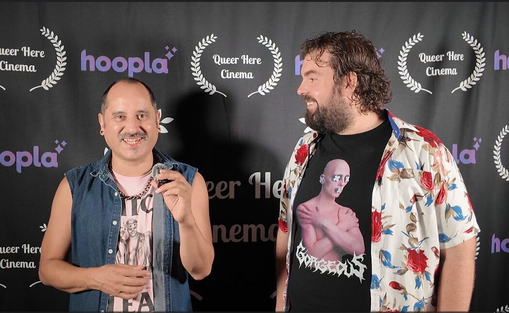 Two men standing in front of a backdrop with logos, smiling and talking, one holding a drink.