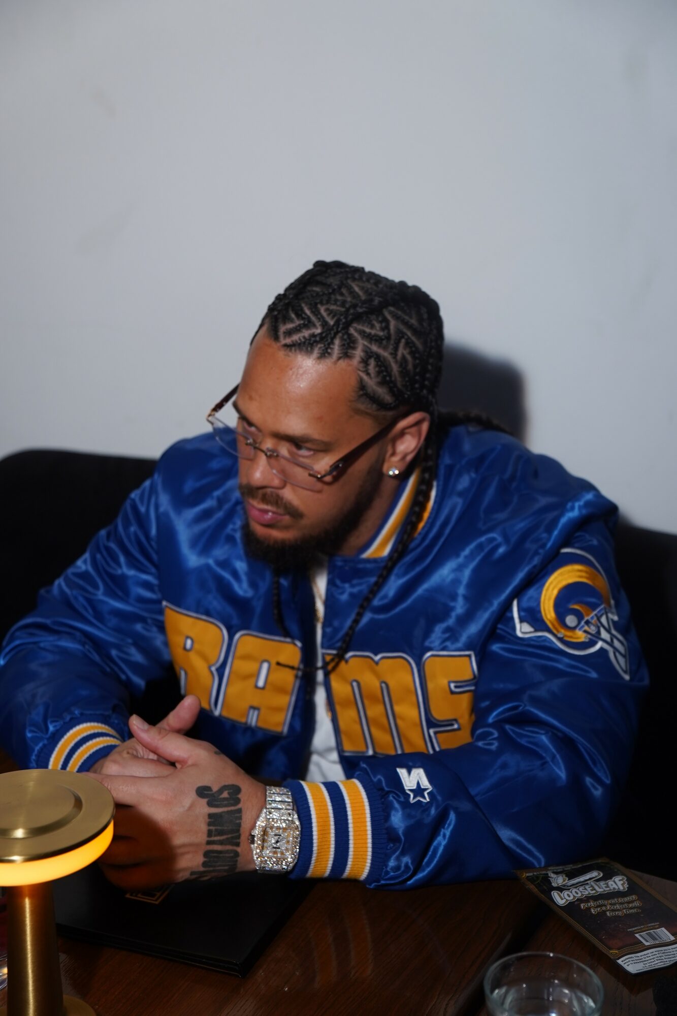 Man with glasses and braided hair wearing a blue Rams jacket, sitting at a table with a phone and a glass.